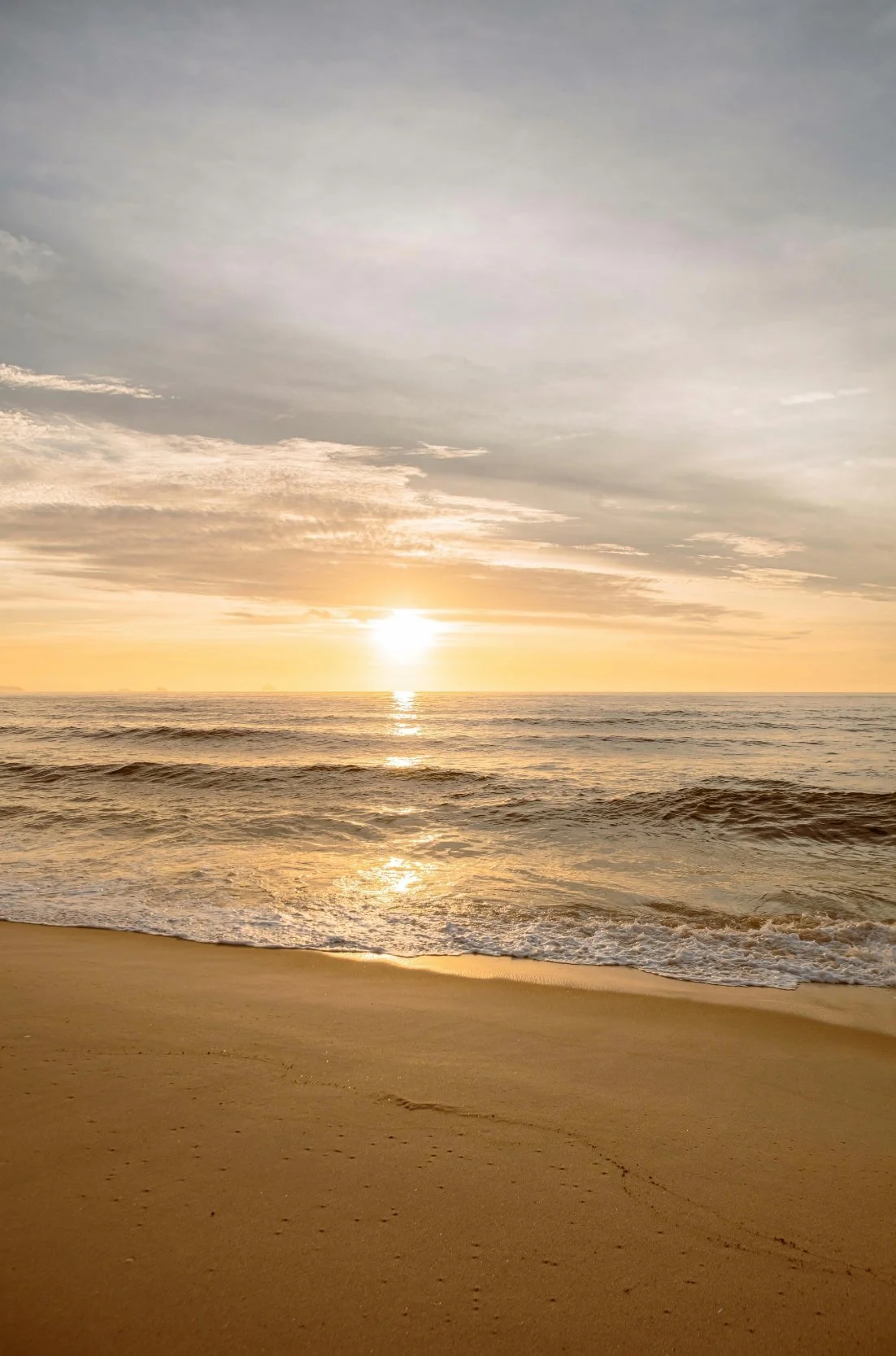 Sunset over the ocean with waves gently hitting the sandy beach, partly cloudy sky.