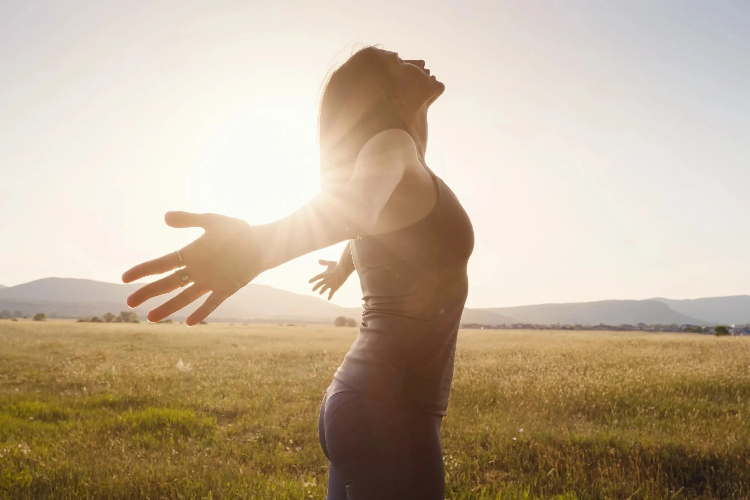 A woman stands in a field with arms outstretched, facing the sun, with a mountain range in the background.