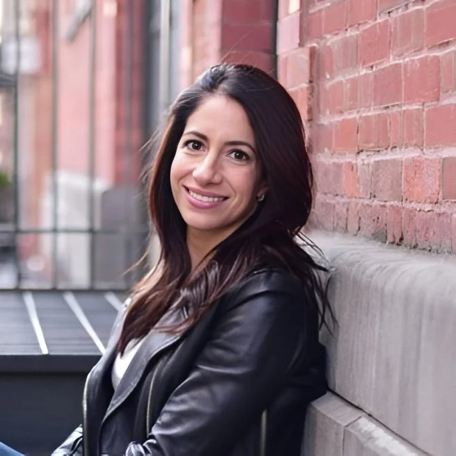 A young woman with long dark hair smiling while sitting on a concrete ledge against a brick wall outdoor setting.