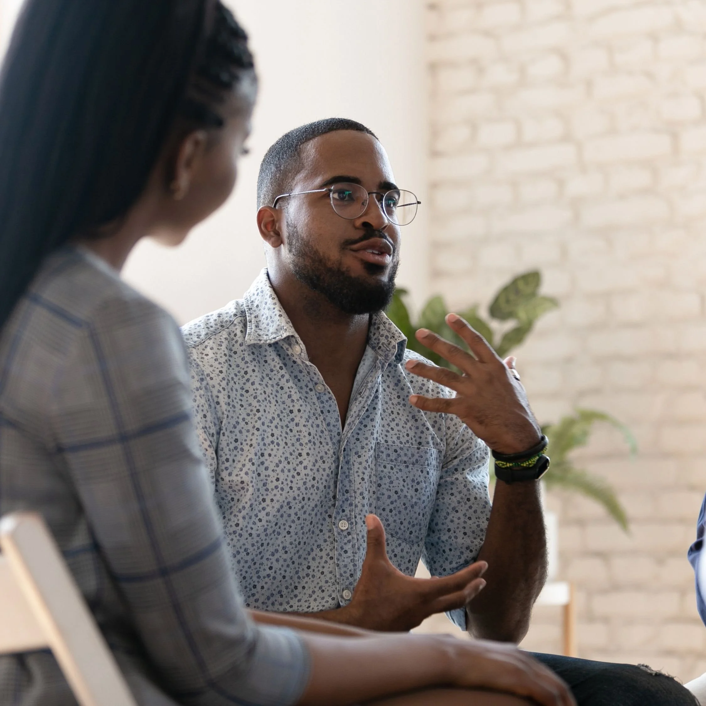 A man with glasses speaking during a discussion in a casual office setting, with a woman listening.