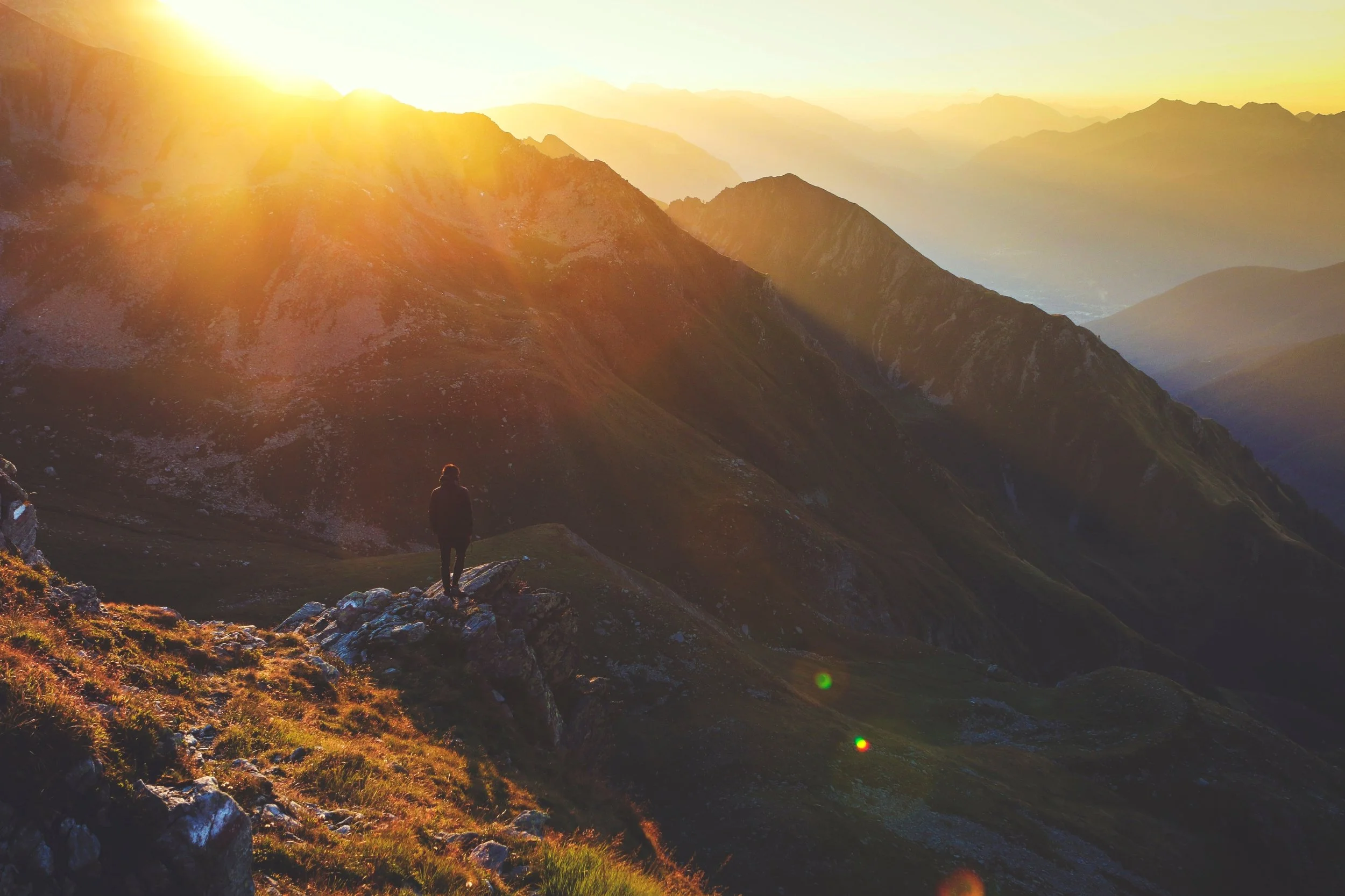 A person standing on a rocky ledge overlooking a mountain range at sunset with sun rays illuminating the sky.