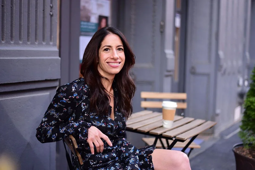 A woman with long dark hair and a black floral dress sitting outdoors at a small wooden table with a coffee cup, smiling at the camera.