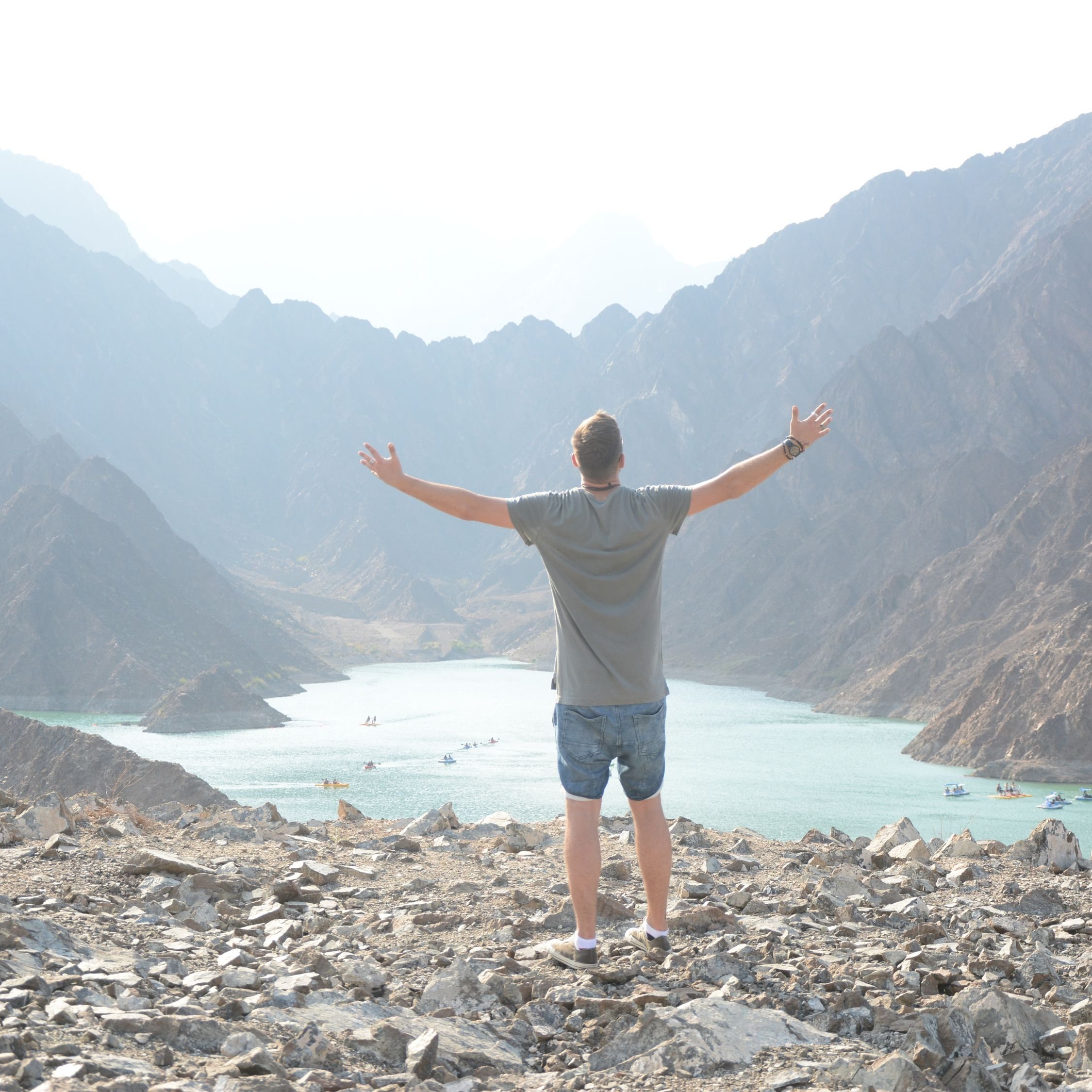 A man standing with arms outstretched on rocky terrain overlooking a lake surrounded by mountains.