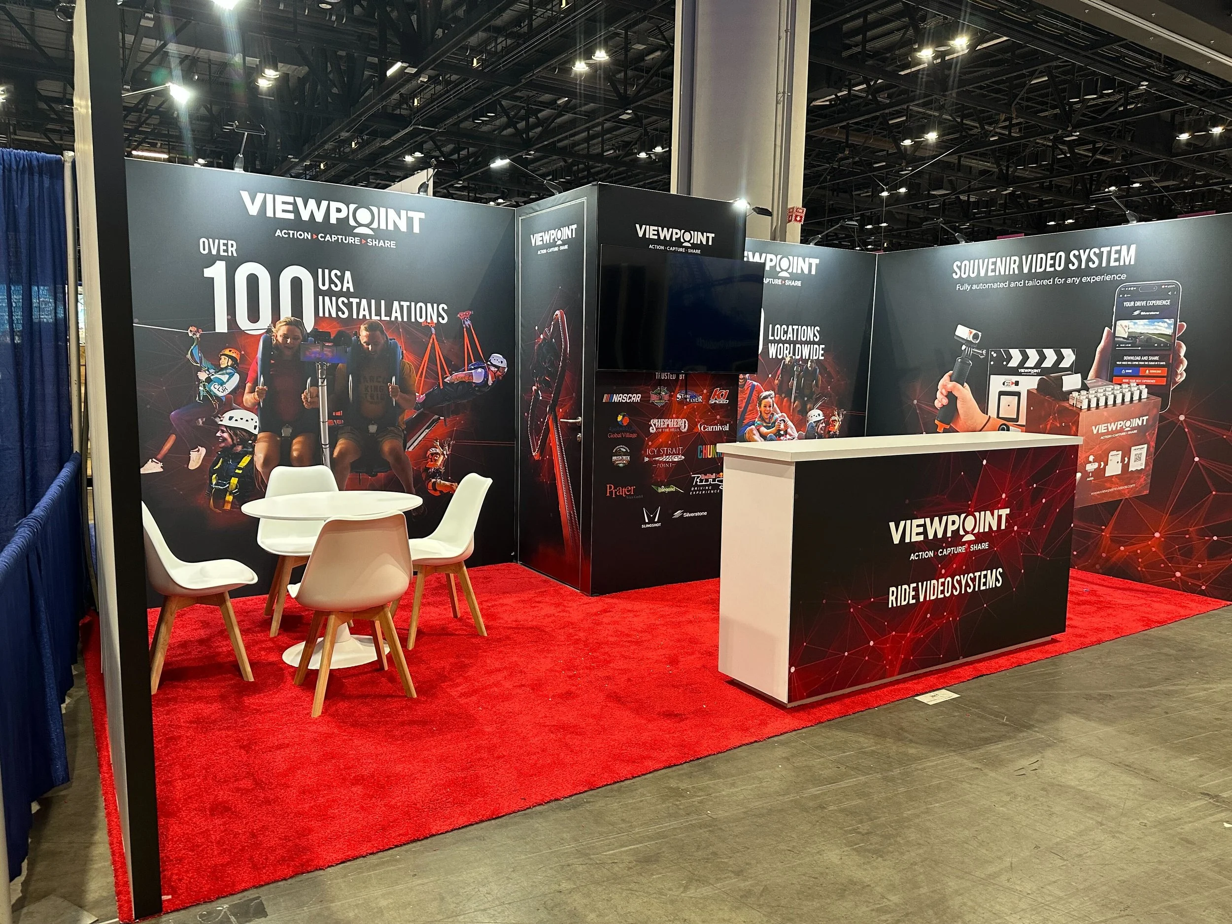 Exhibition booth for ViewPoint, showcasing video system products. The booth has large graphics with people biking, skiing, and snowboarding. Signs highlight over 100 USA installations and worldwide locations. There are chairs and a small table with a bright red carpet on the floor.
