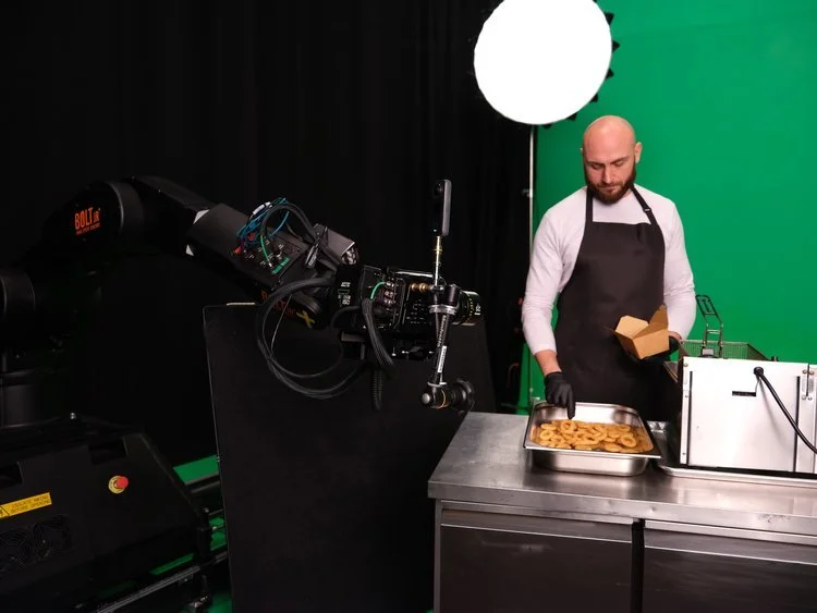 A man wearing a black apron and gloves is doing a cooking segment in front of a green screen and professional camera, preparing cookies.