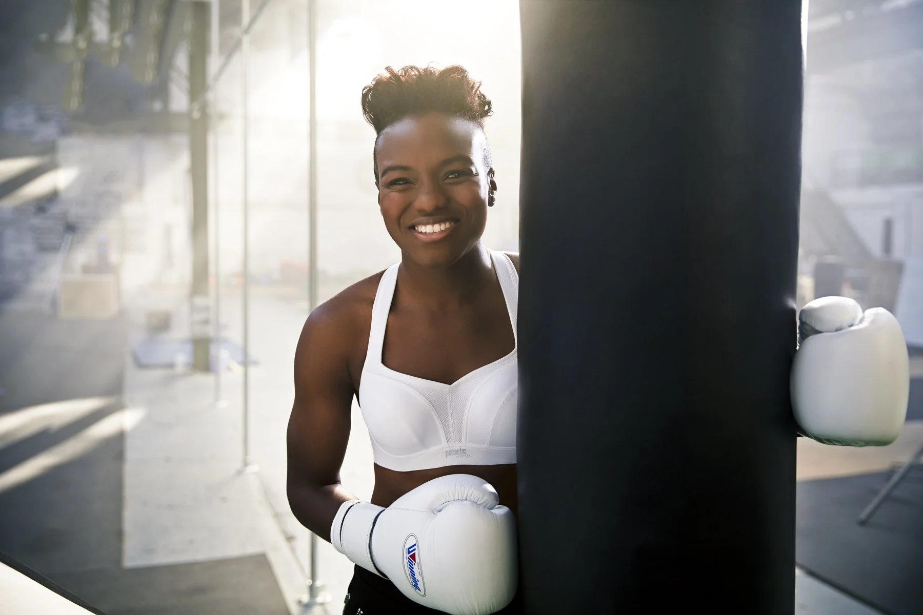 A smiling Nicola Adams boxer with short curly hair, wearing a white sports bra and boxing gloves, practicing her punches on a black punching bag in a bright gym.
