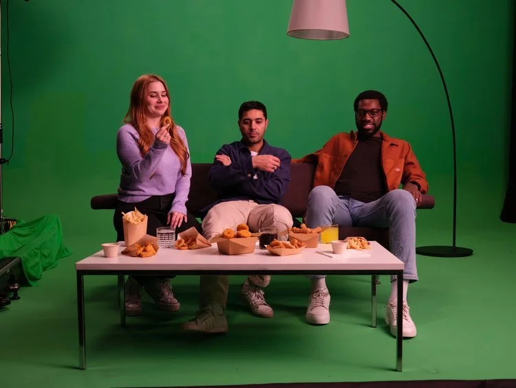 Three people sitting on a sofa in front of a green screen with food and drinks on a table in front of them during a filming or photo shoot.
