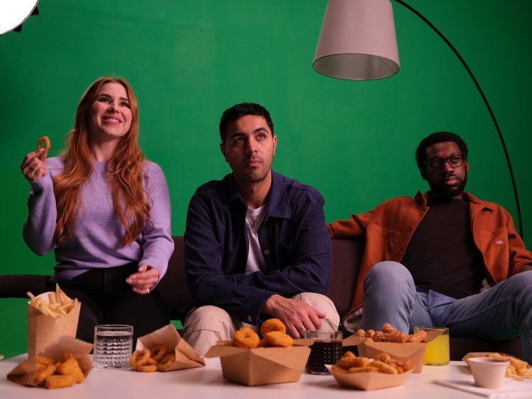 Three friends sitting on a couch in front of a green screen, with snacks and drinks on the table in front of them, watching something with varied reactions.