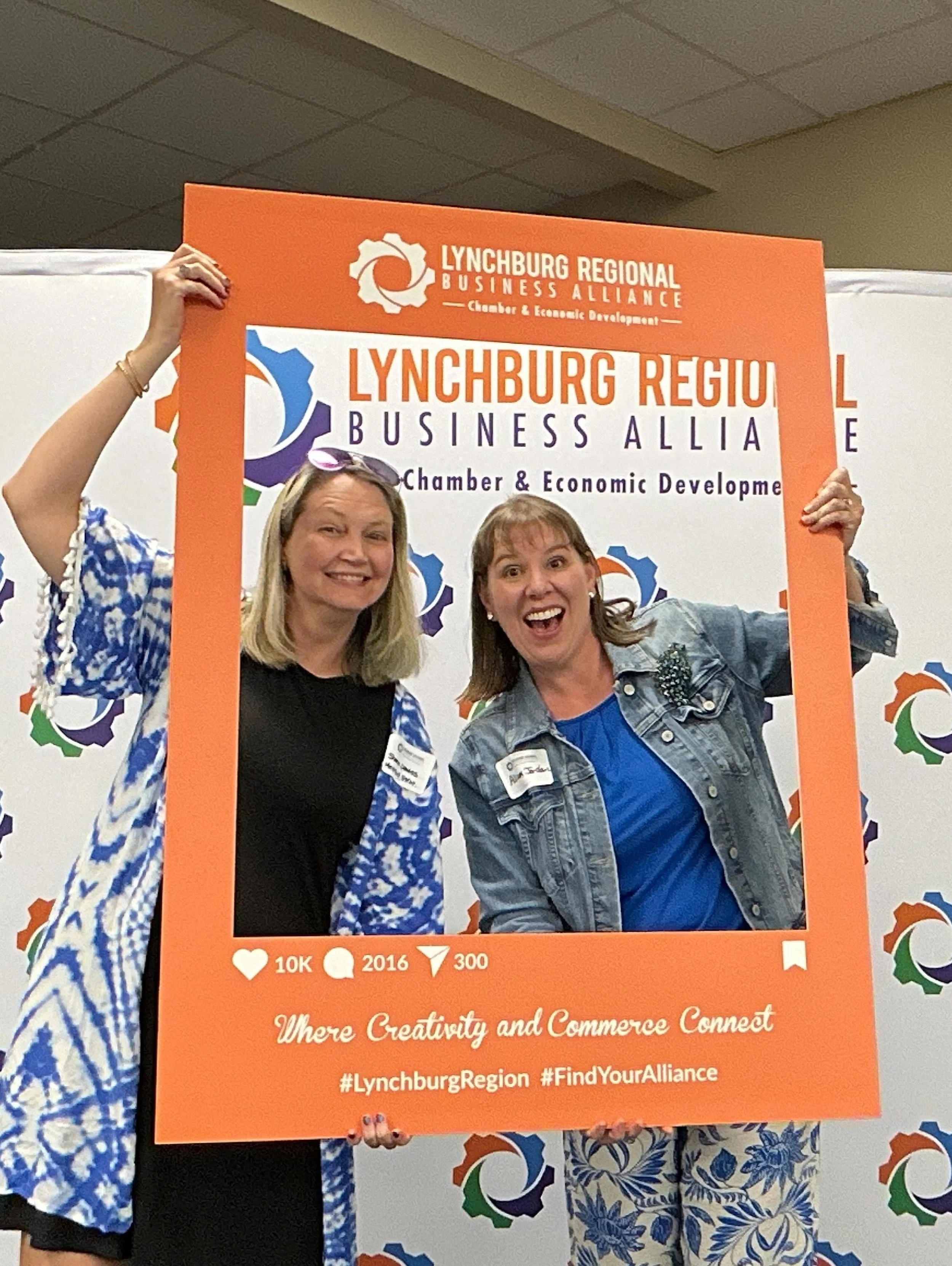 People holding up an orange sign with the Lynchburg Regional Business Alliance logo and slogan, with a background featuring colorful gears and the text 'Where Creativity and Commerce Connect'.