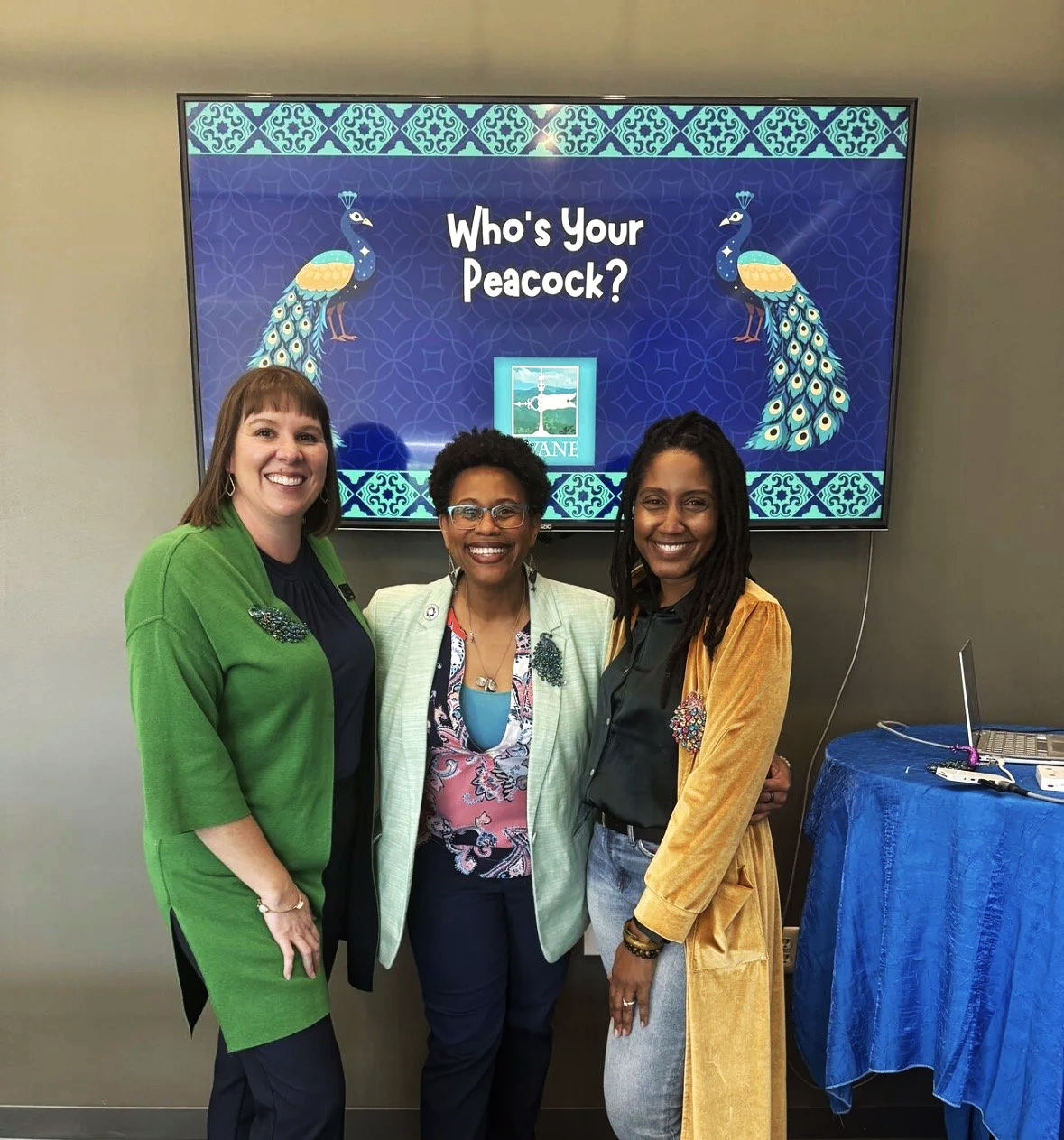 Three women smiling in front of a digital display that says "Who's Your Peacock?" with a peacock graphic and the text "LANE" beneath it, standing indoors with a table to the right covered in blue cloth and electronic equipment.