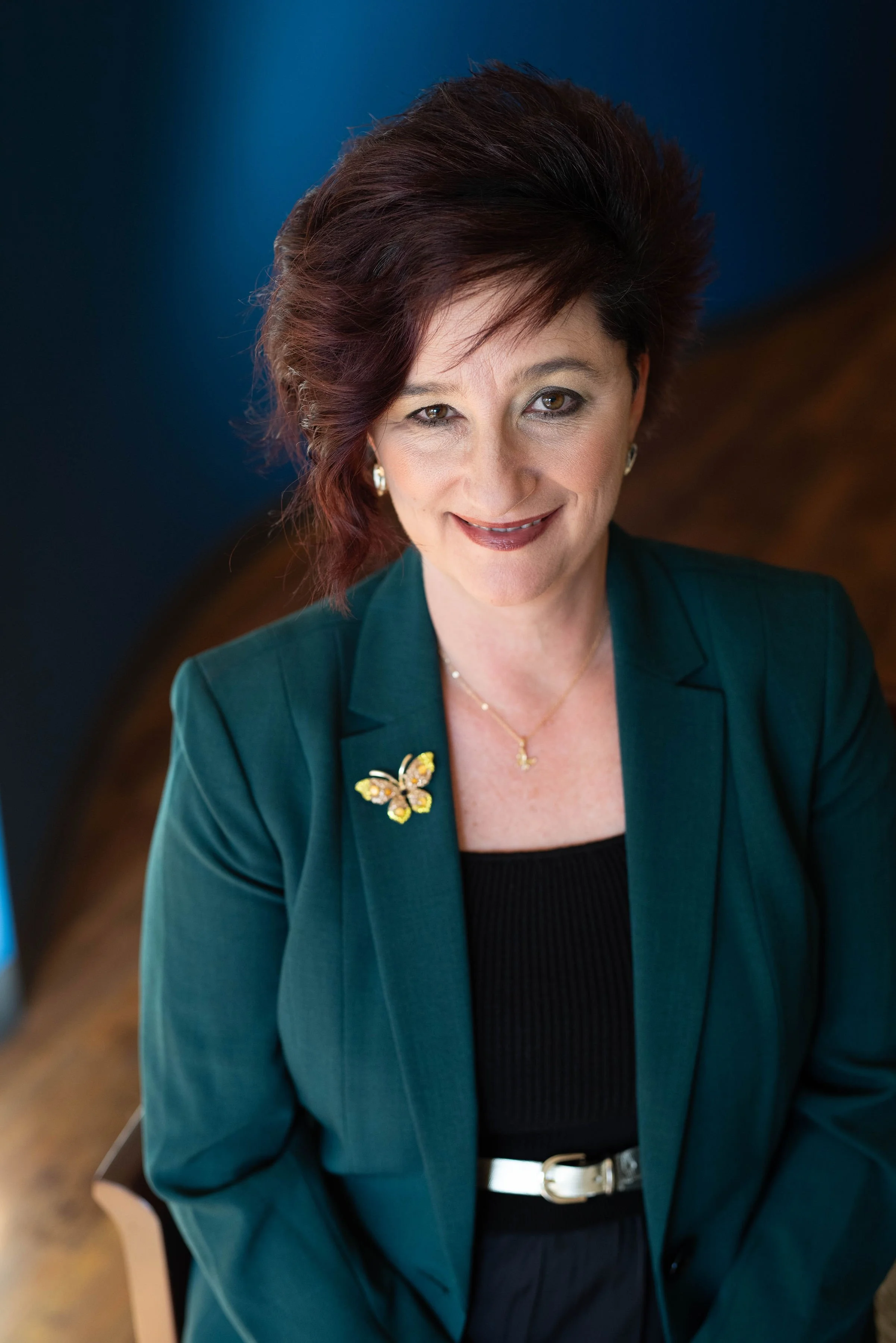 A woman with short, dark red hair and light makeup, wearing a dark green blazer with a butterfly pin, smiling with one eye closed, seated in an indoor setting with a blurred background.