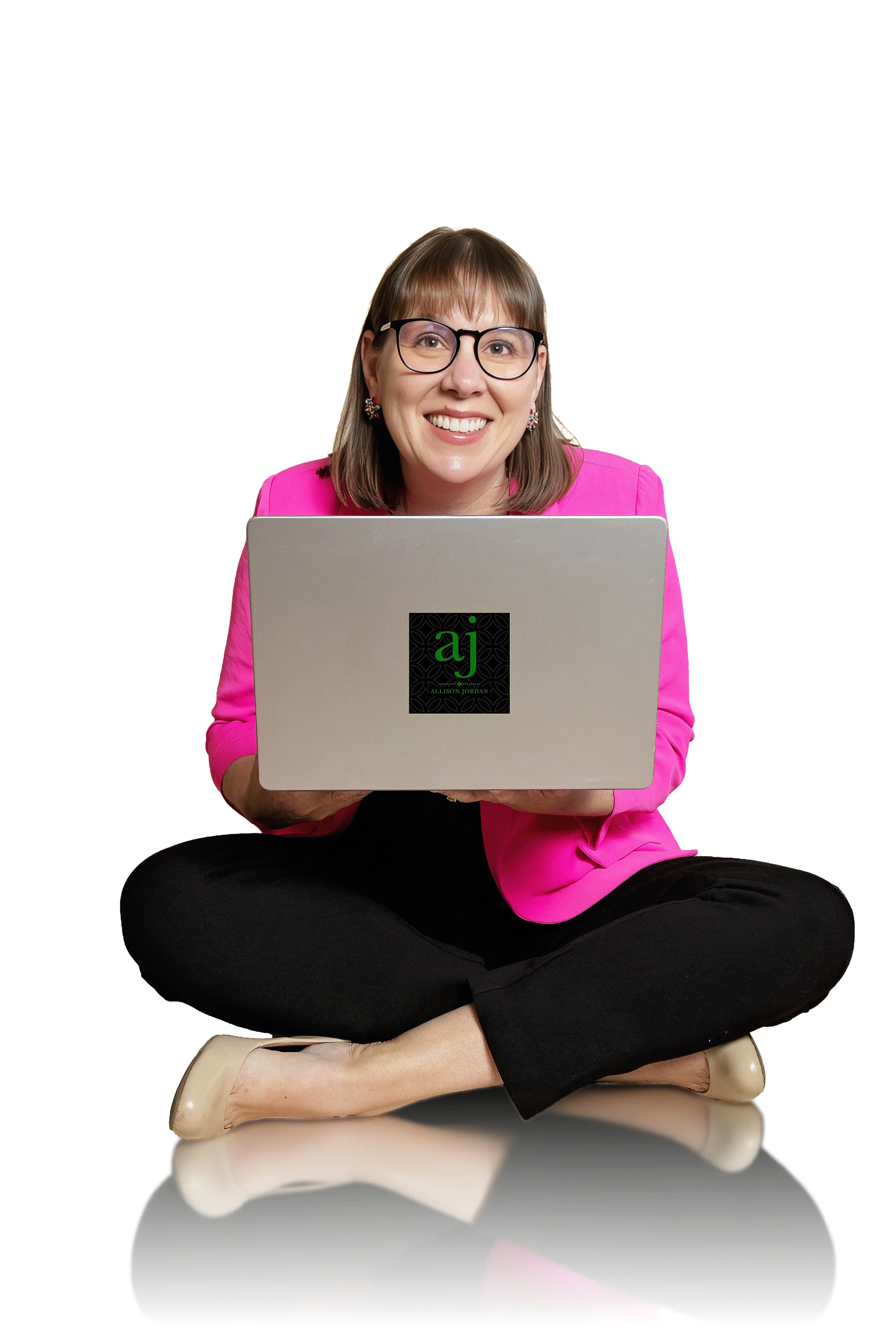A woman with brown hair, glasses, and earrings, wearing a pink blazer and black pants, sitting cross-legged on a beige chair, smiling, and holding a silver laptop with a black sticker that has green initials 'aj' and her name Alison Jordan, against a black background.