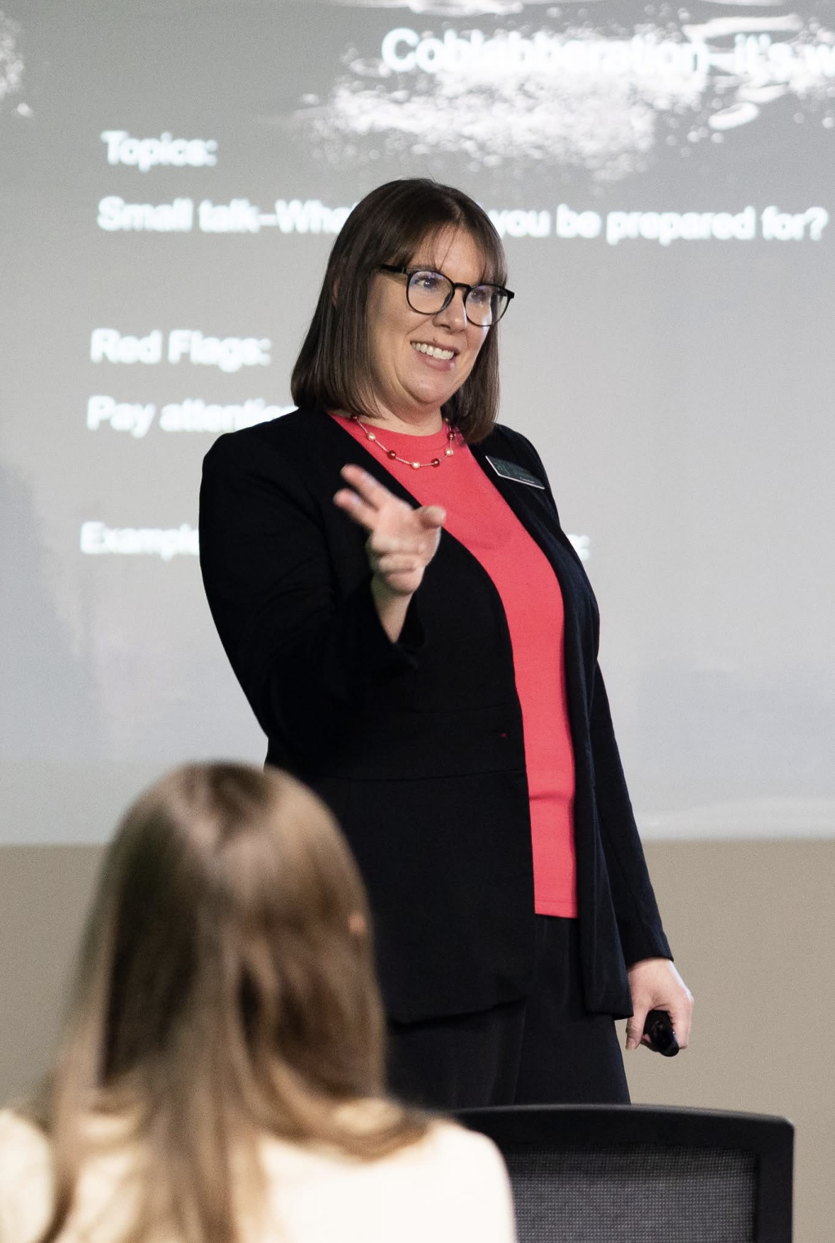 A woman with glasses, wearing a black blazer over a red top, smiling and gesturing towards the audience during a presentation. In the background, a slide with topic information is visible.