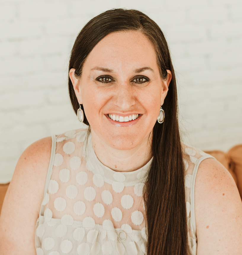 A woman with long brown hair and earrings, smiling, wearing a sleeveless cream-colored top with a dotted pattern, against a white brick wall background.