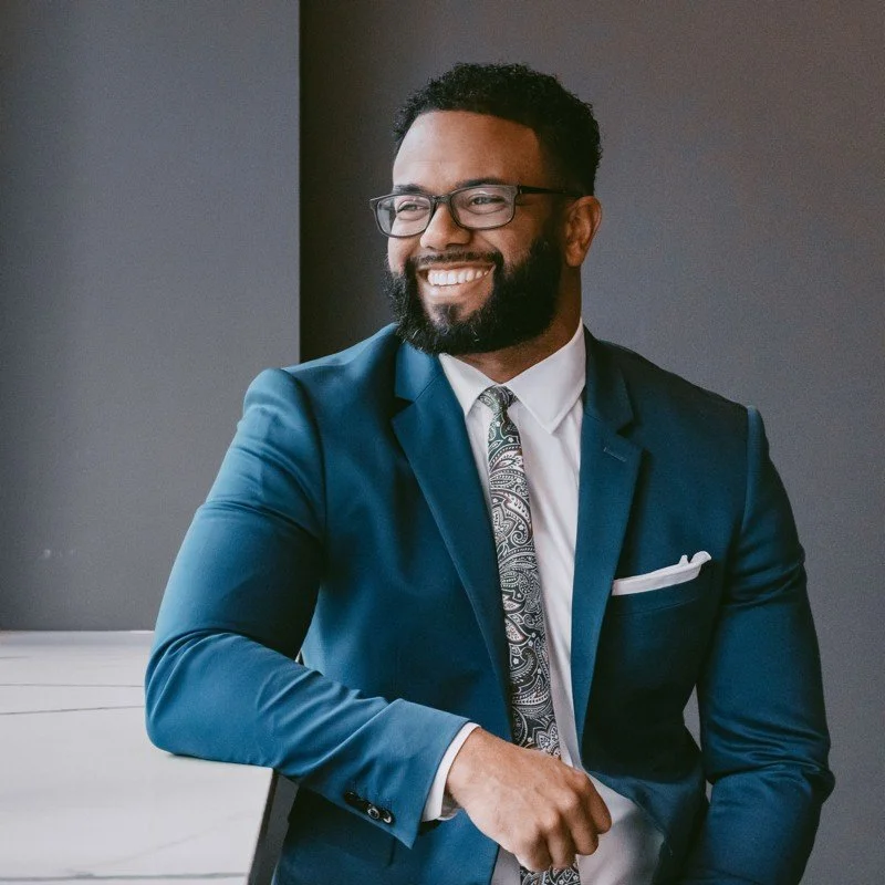 A smiling man in a blue suit, white shirt, patterned tie, and glasses, standing indoors against a gray background.