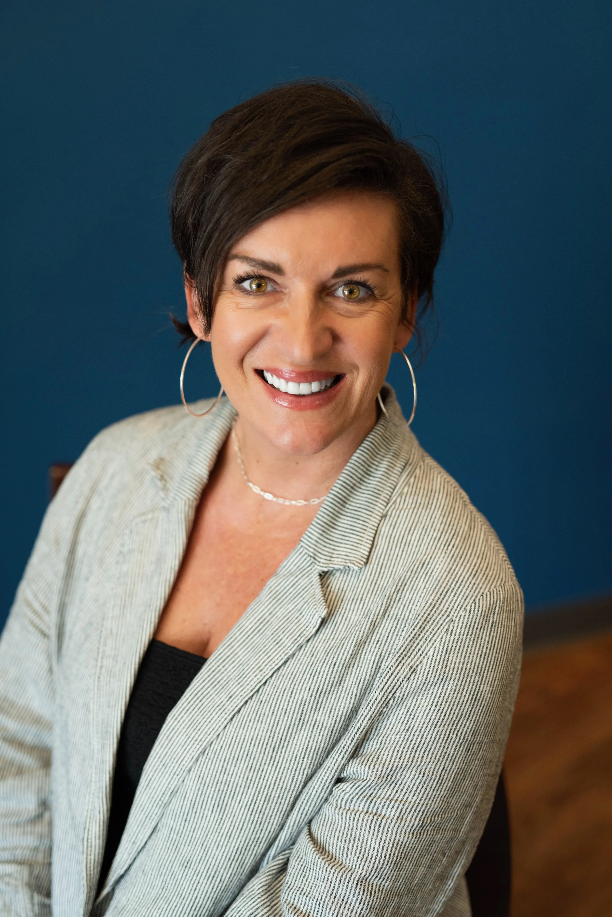 A woman with short dark hair, wearing hoop earrings, a light-colored blazer, and a black top, smiling and looking at the camera against a dark blue background.