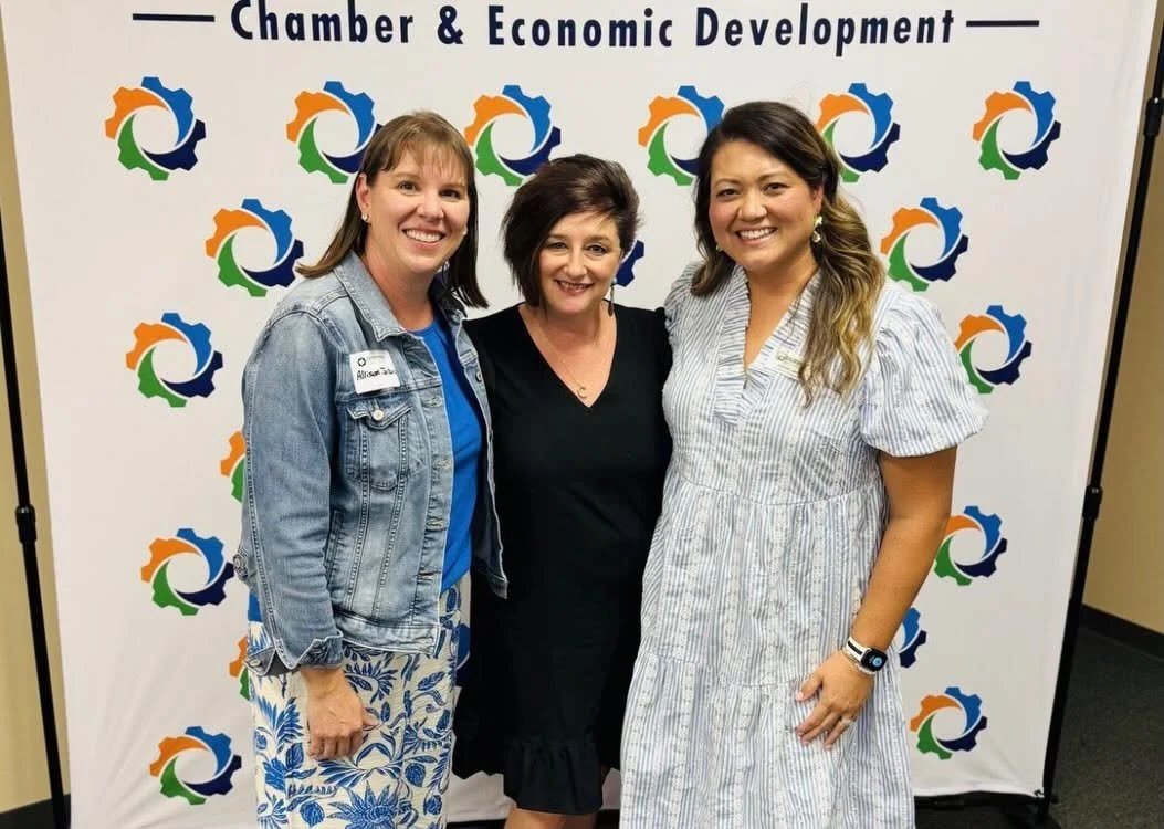 Three women standing together, smiling, in front of a backdrop with the text 'Chamber & Economic Development' and a colorful gear logo repeating across it.