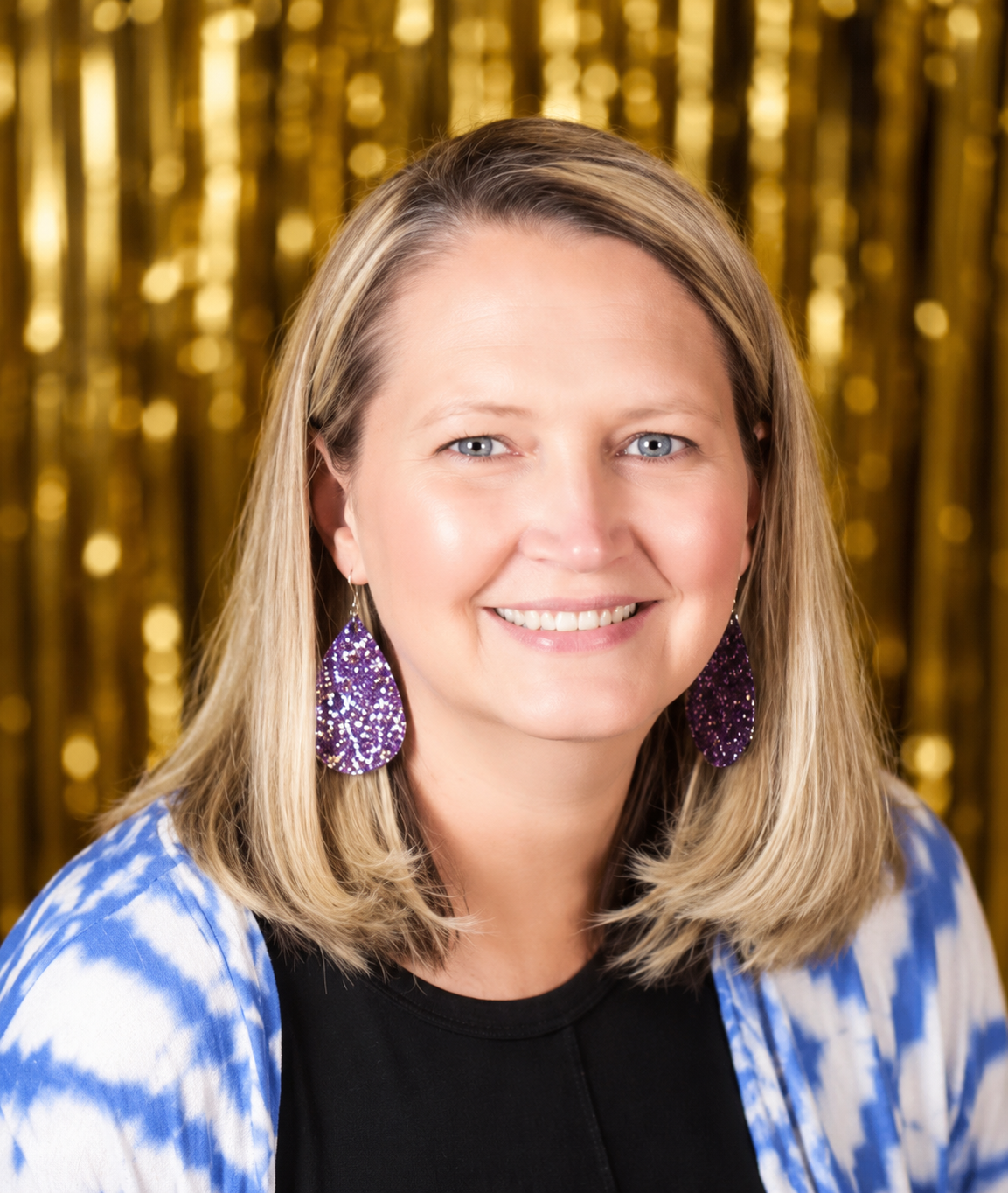 A woman with blonde hair, wearing purple glittery earrings, smiling in front of a gold shimmering background, dressed in a black top and a blue and white tie-dye patterned sweater.