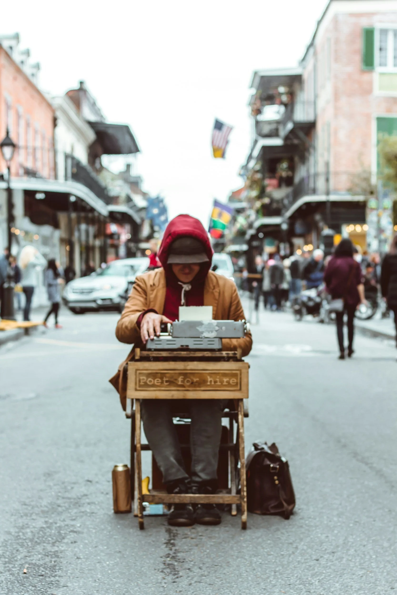 Street performer dressed as a poet typing at a vintage typewriter on a wooden cart with a sign that reads 'Poet for hire' during daytime.