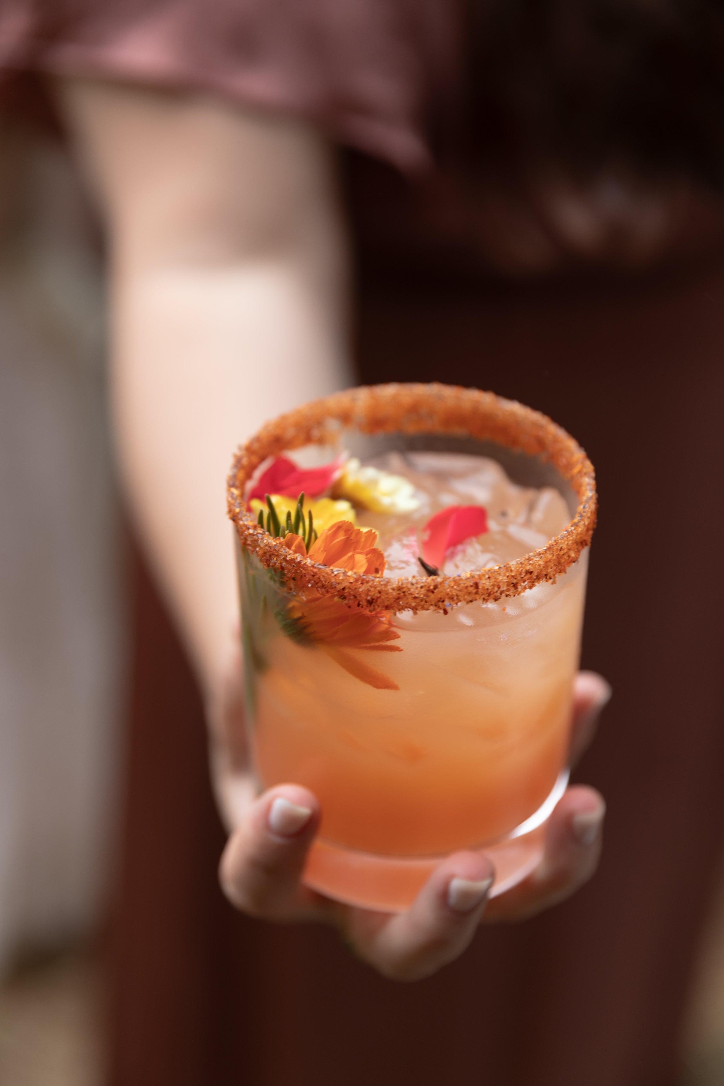 A person holding a cocktail with a salted rim, garnished with flowers, and ice cubes in a glass.