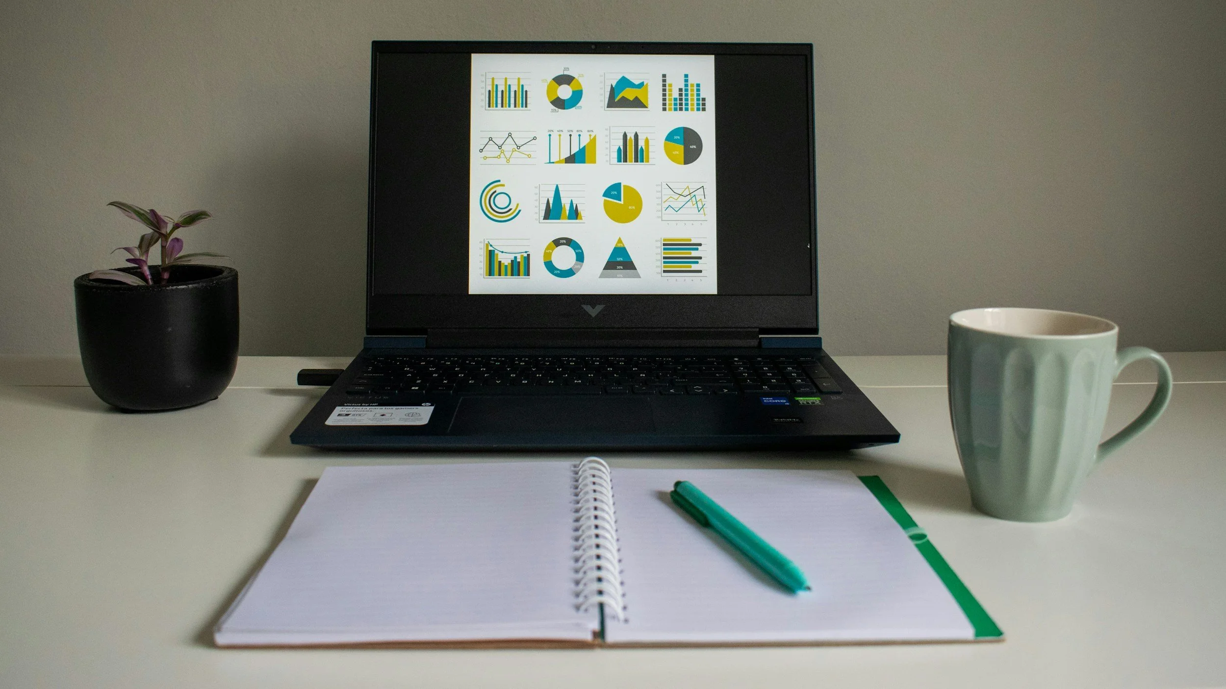 A desk setup with an open notebook and green pen, a black laptop displaying colorful charts and graphs, a potted plant on the left, and a white mug on the right, all on a white surface against a gray wall.