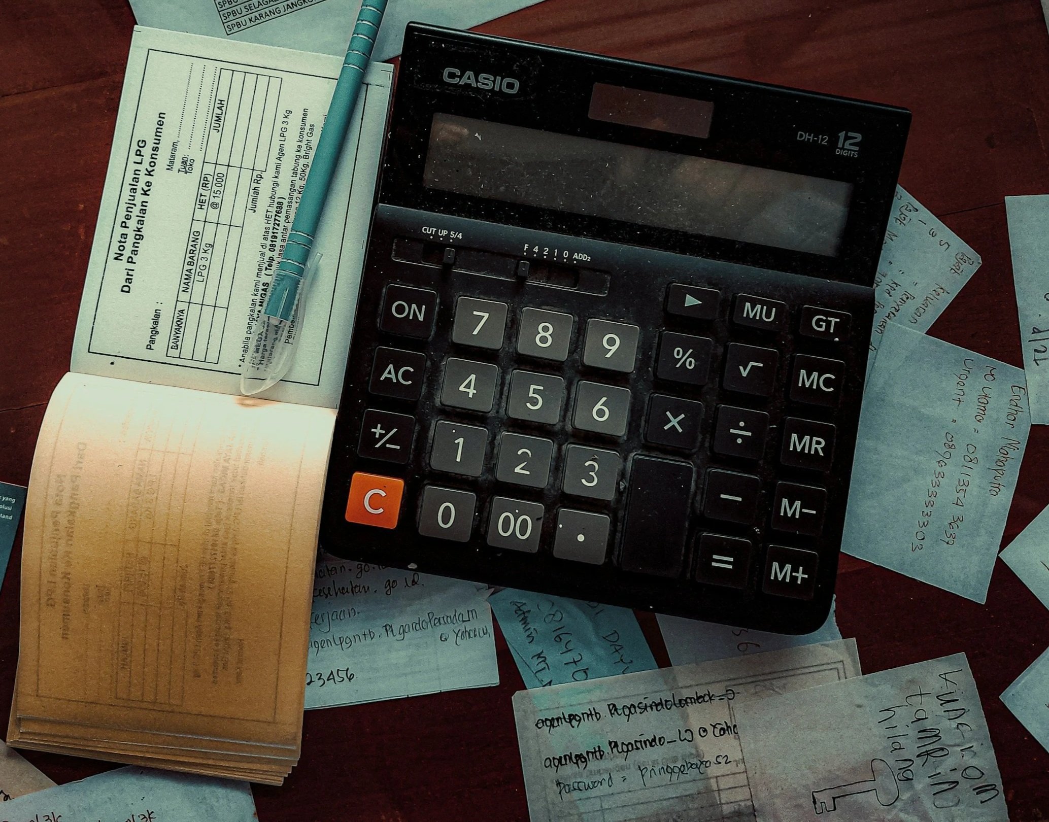 A black calculator, a stack of yellow receipts, a blue pen, and several handwritten notes are spread on a wooden surface.