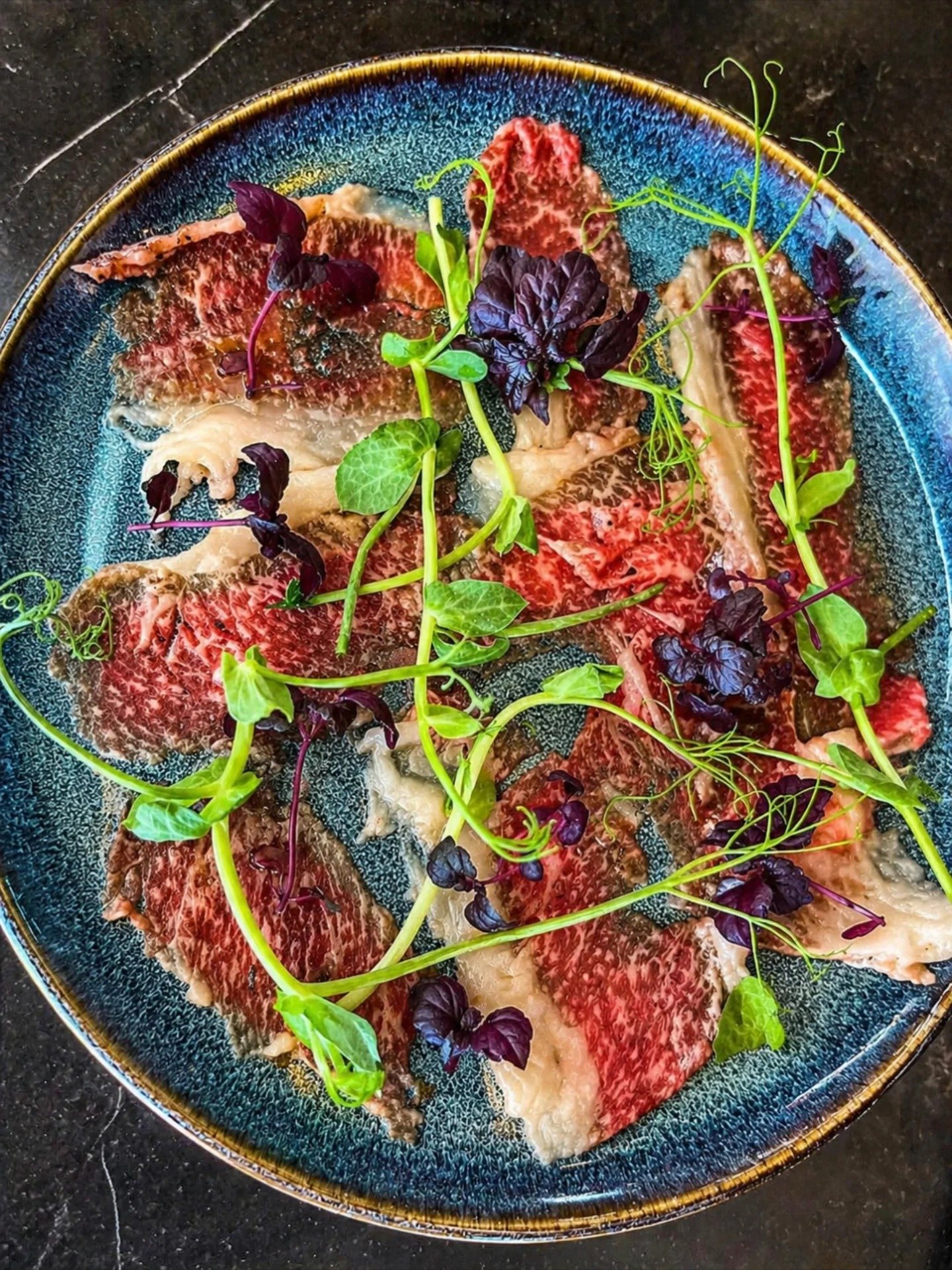 Wagyu Carpaccio –  Wagyu brisket tynt skivet med sesamolje, urter og små grønne spirer på en blå keramisk tallerken. - Rolis Bodega, Nordstrand, Oslo