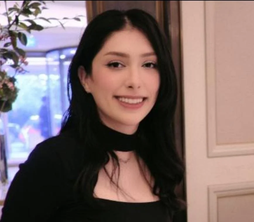 A young woman with long black hair smiling indoors, with a window and some greenery in the background.