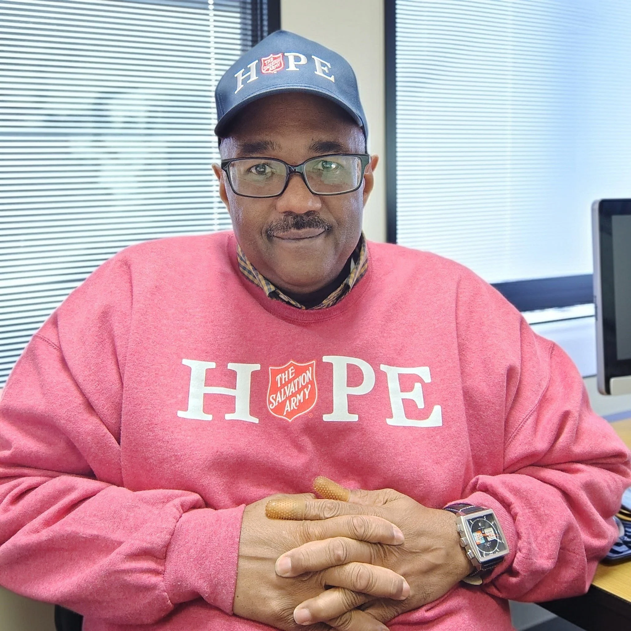 Close-up of a person wearing glasses and a pink sweatshirt with the Salvation Army logo, sitting at a desk with a window with blinds in the background.