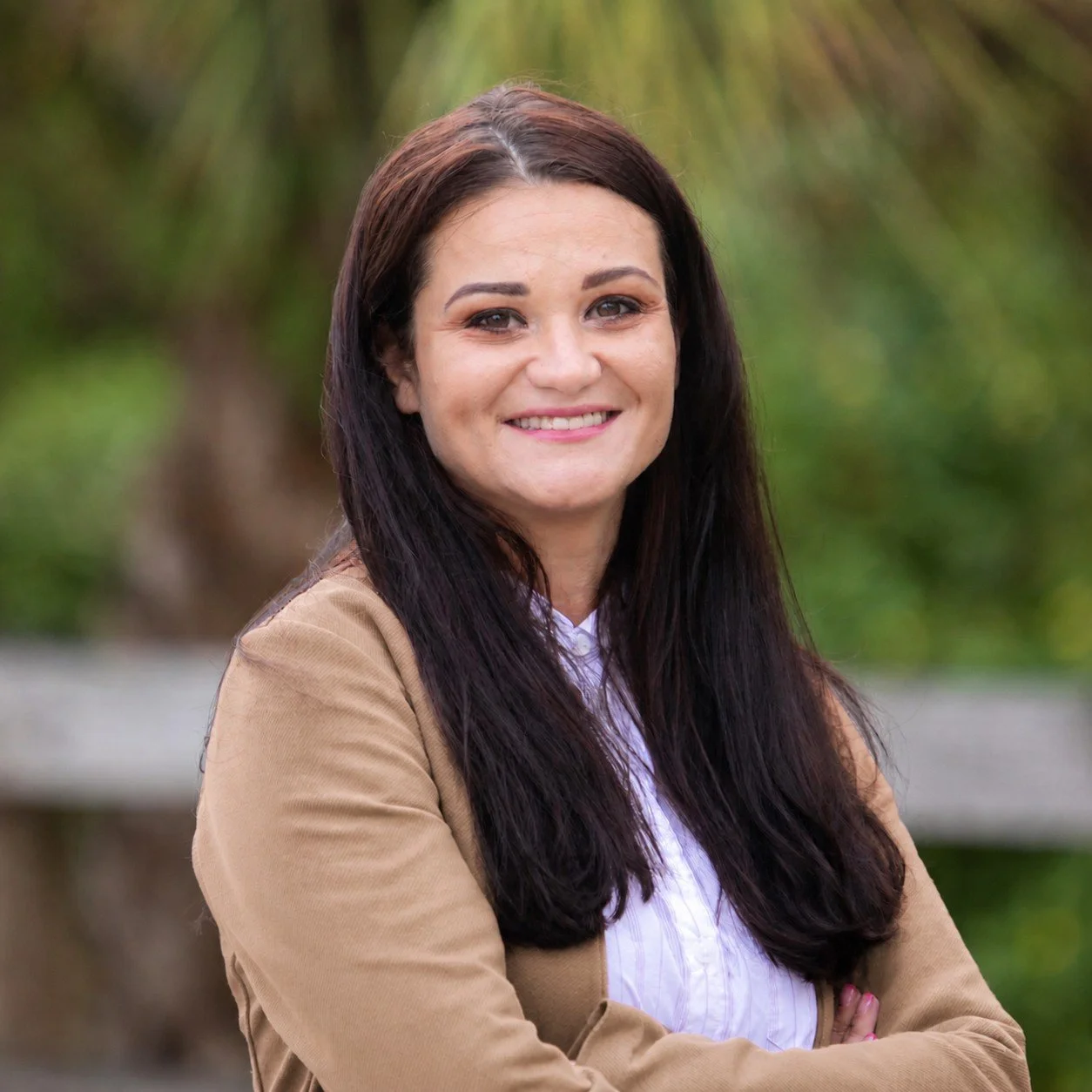 A woman with long dark hair smiling outdoors in front of blurred greenery and a wooden fence.
