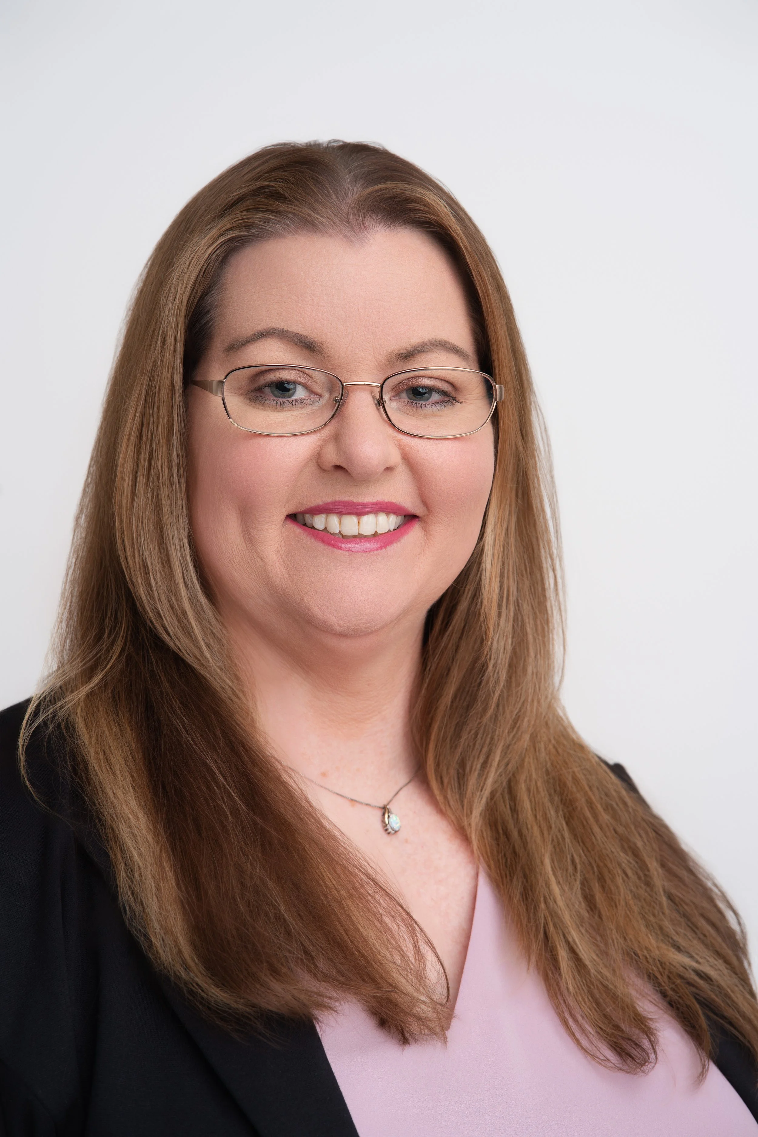 Close-up of a woman with long brown hair, wearing glasses, a light pink top, and a black blazer, smiling against a plain white background.