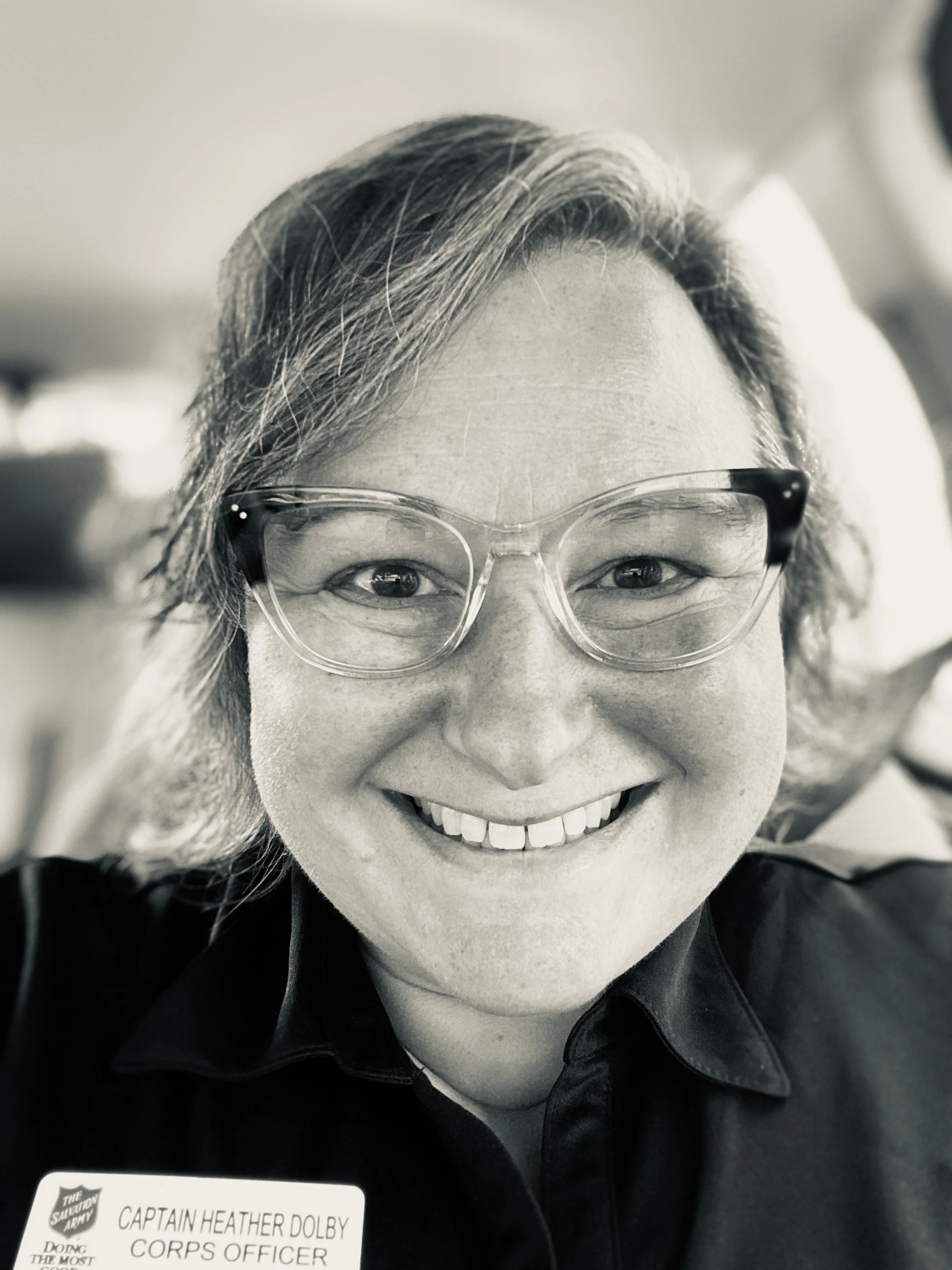 Close-up of a woman with glasses smiling inside a vehicle, with a name tag that reads 'Captain Heather Dolby, Corps Officer'.