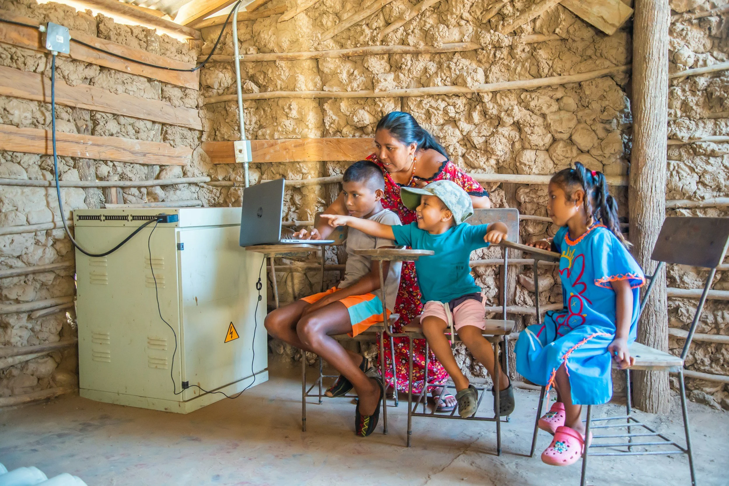 A woman, likely a teacher or caretaker, helps three children with a laptop inside a rustic, earthen-walled room. The children are engaged, with one pointing at the screen. The space has exposed wooden beams and wiring, and the floor appears to be concrete or packed earth.