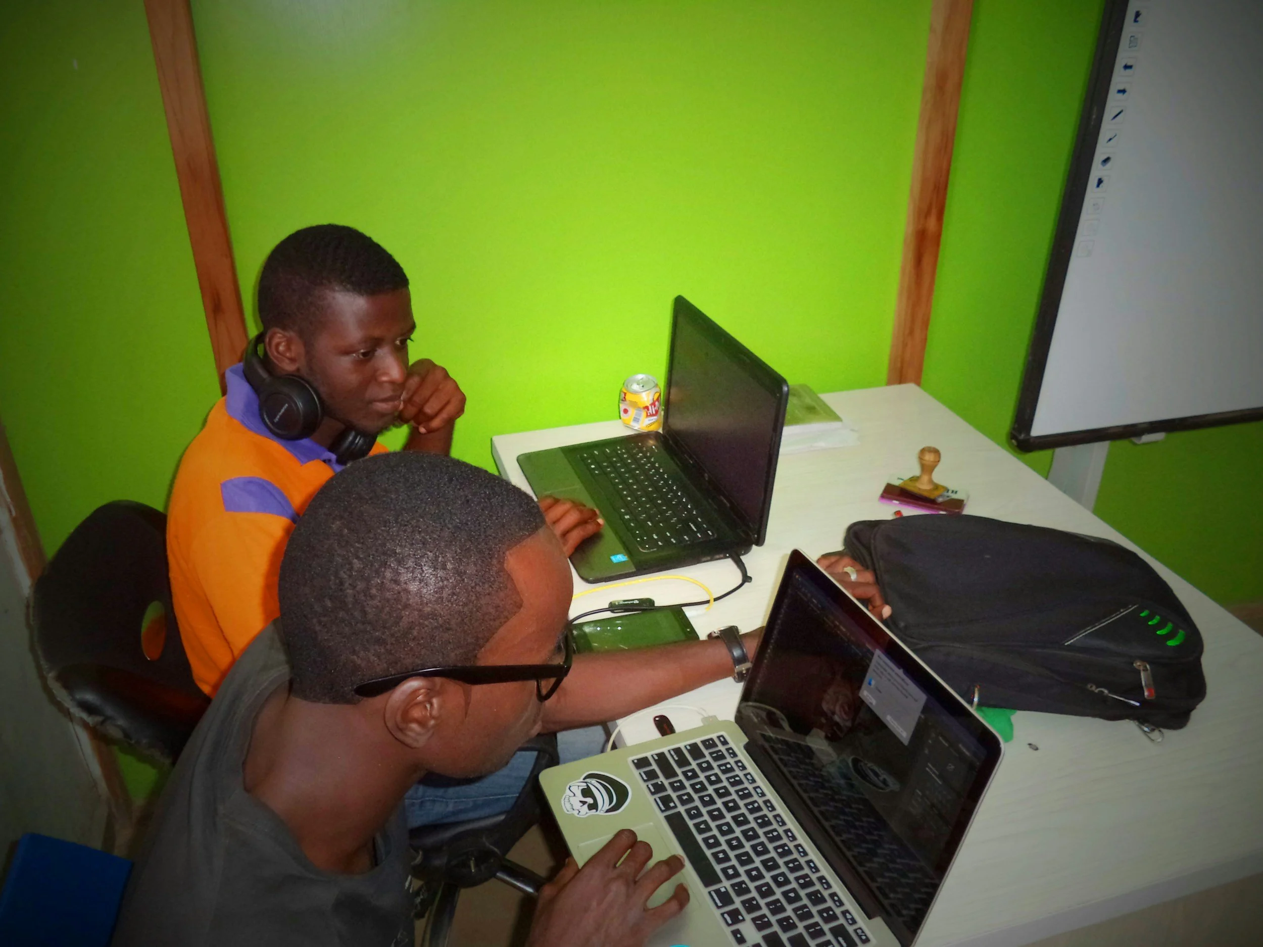 Two men working on laptops at a desk, one with headphones and the other with glasses, in a room with bright green walls.