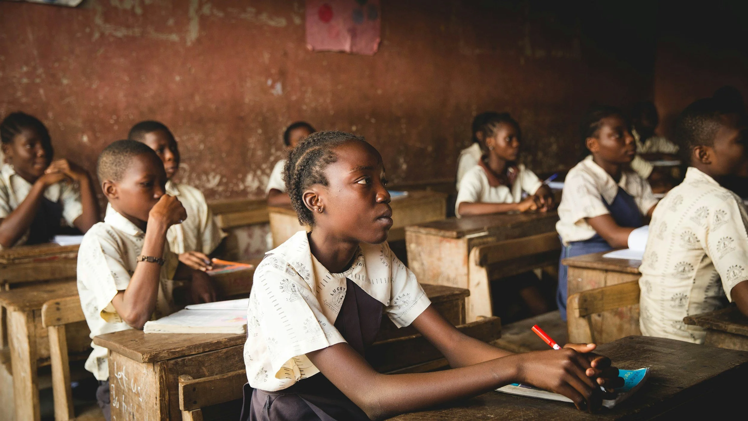 A classroom with young students sitting at wooden desks, attentively listening, with notebooks and pens in an educational setting.