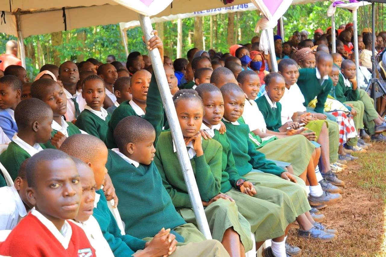 A large group of school children and teachers seated under a tent outdoors surrounded by greenery. The children wear green and white school uniforms and appear attentive to an event.