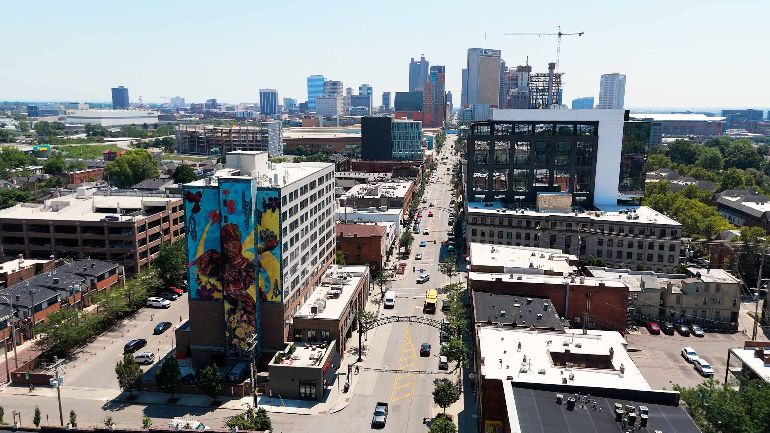 View of downtown cityscape with tall buildings, streets, cars, and trees under a clear blue sky.