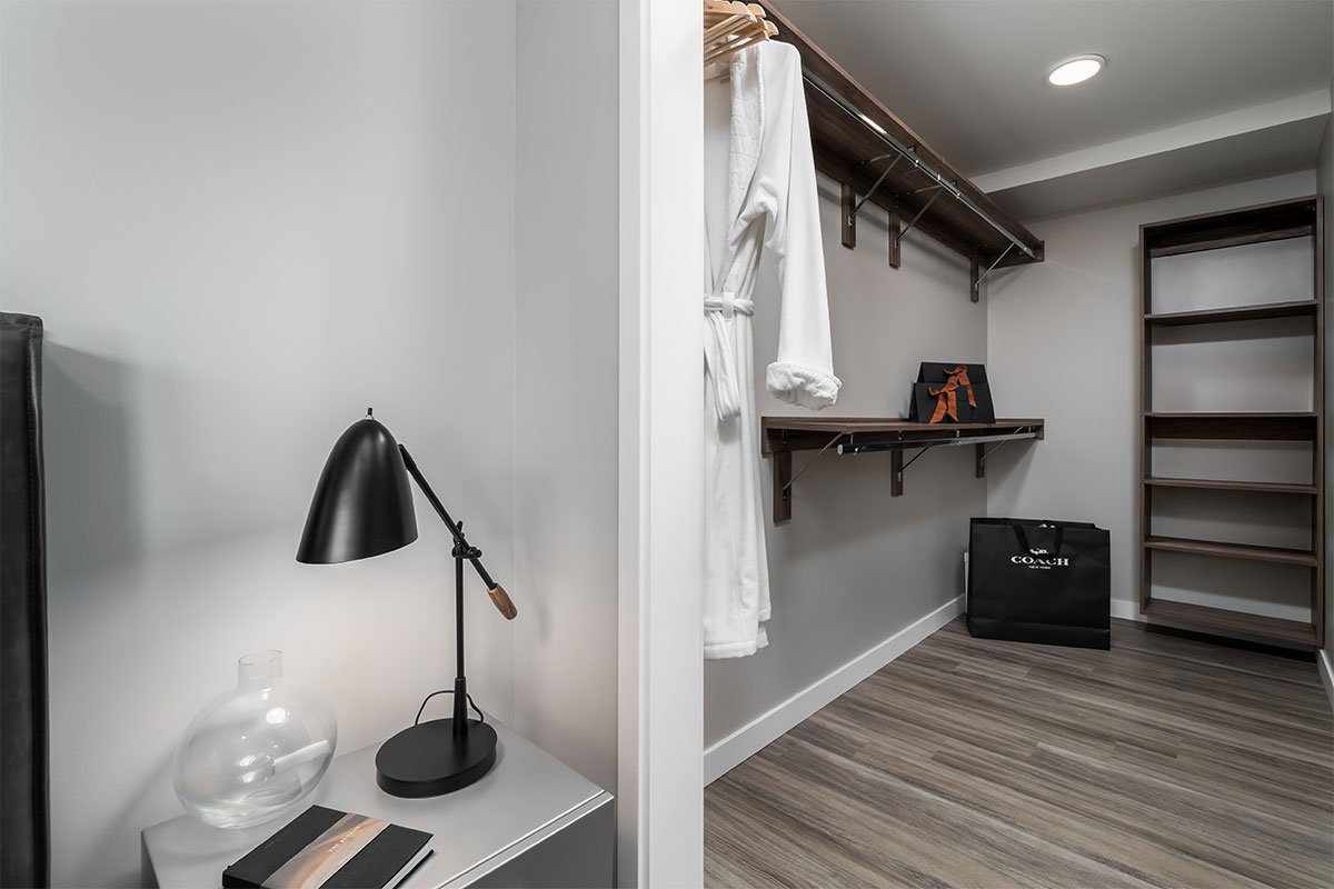 A walk-in closet with gray walls, wooden shelves, and a hanging white bathrobe. A black Coach shopping bag is on the floor, and a black box with a bow is on a wall shelf. The floor has wood grain pattern, and a ceiling light illuminates the space.