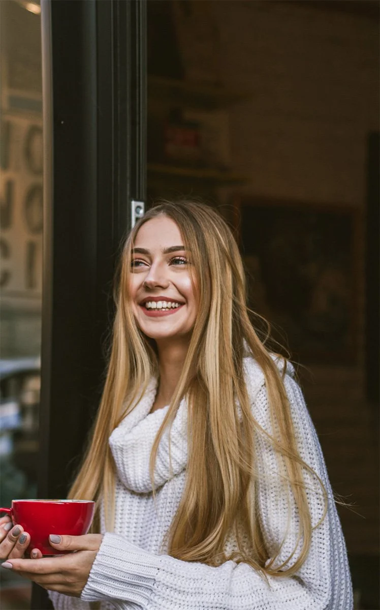A young woman with long blonde hair smiling and holding a red mug, standing near a doorway in an indoor setting.