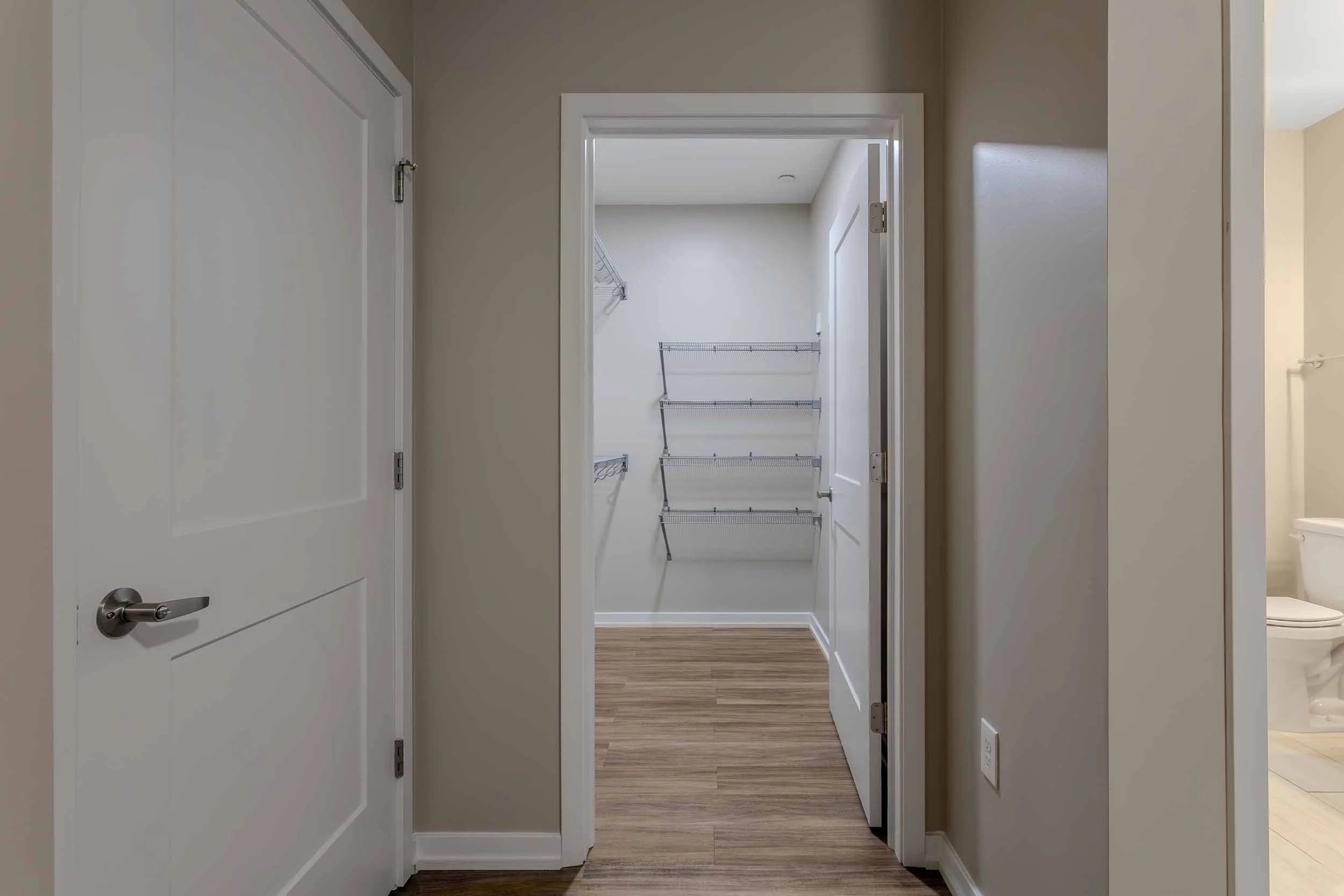 Empty walk-in closet with wire shelves and natural light, hardwood flooring, adjacent bathroom with toilet visible.