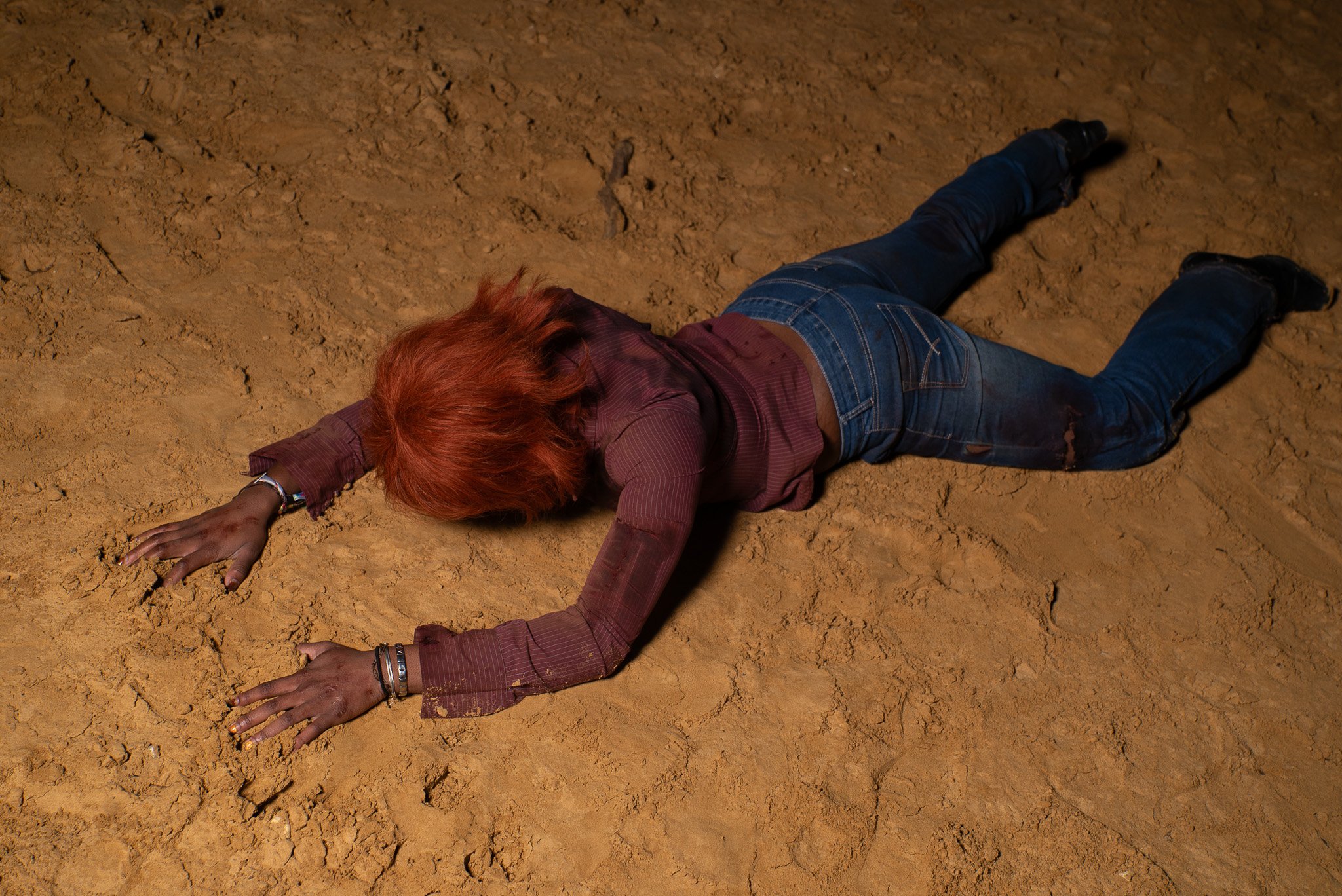 A person with red hair wearing a maroon long sleeve shirt and blue jeans is lying face down on cracked, dry, orange dirt with arms extended forward.