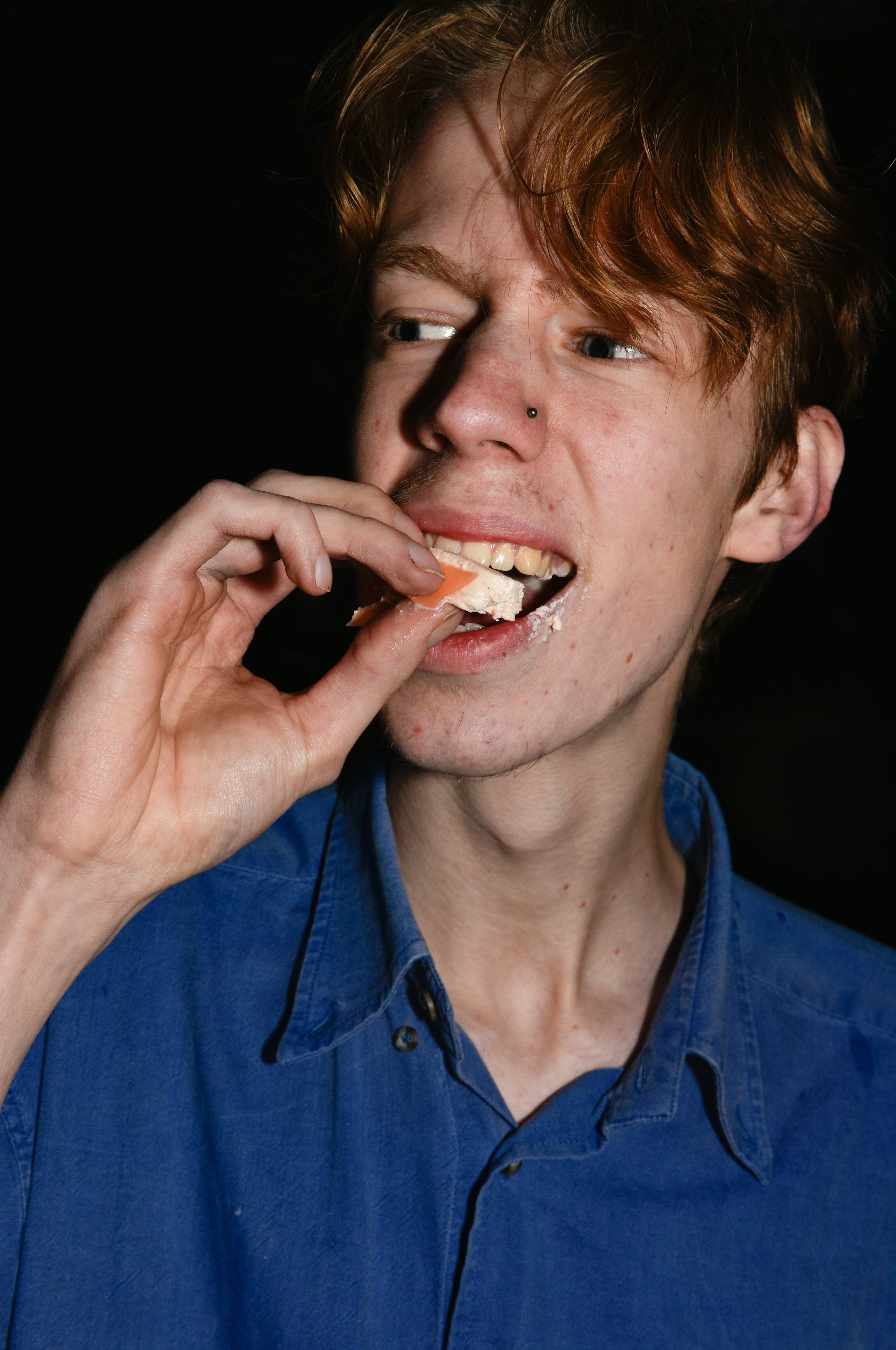 Young man with red hair, a septum piercing, and a blue shirt, eating a slice of pizza with some cheese, against a black background.