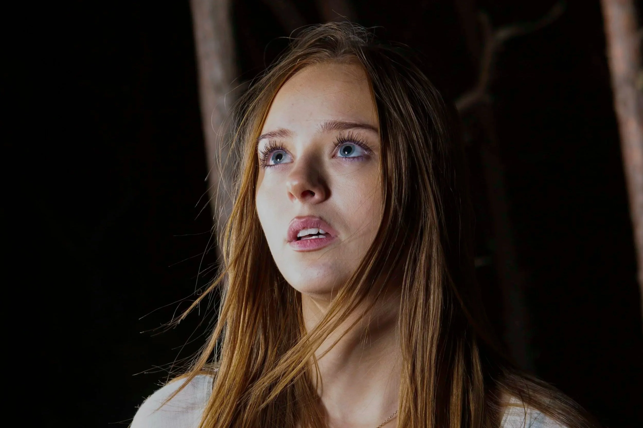 Close-up of a young woman with long, red hair and blue eyes looking upward against a dark background.