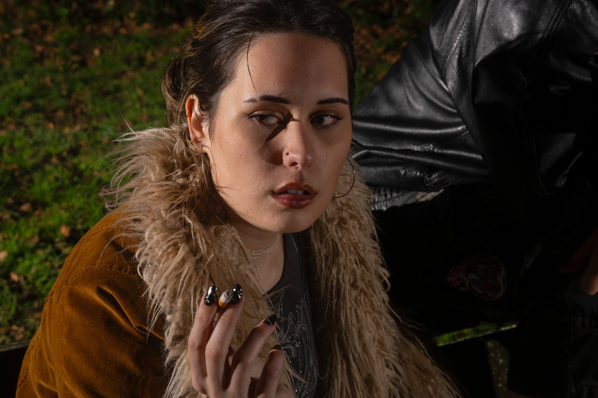 A young woman with dark hair, wearing a fur-lined jacket and makeup, is looking up at the camera while holding a cigarette in her left hand. The background shows grass and leaves, suggesting an outdoor setting.