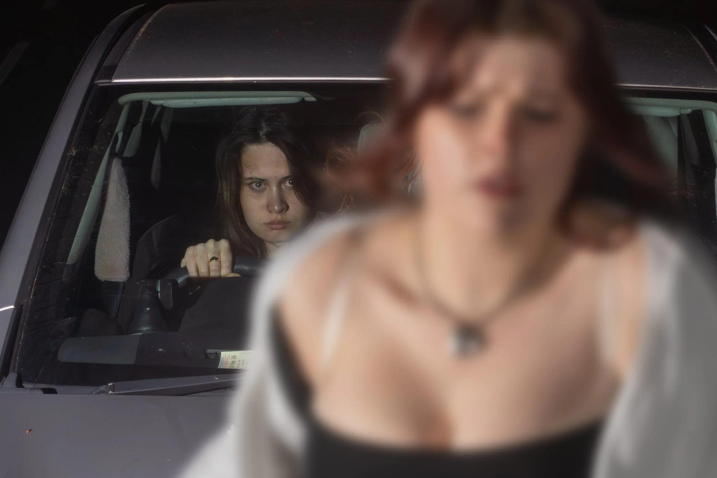 A woman with dark hair sitting inside a vehicle, looking forward, with a serious expression. A person with brown hair and fair skin is blurred in the foreground.
