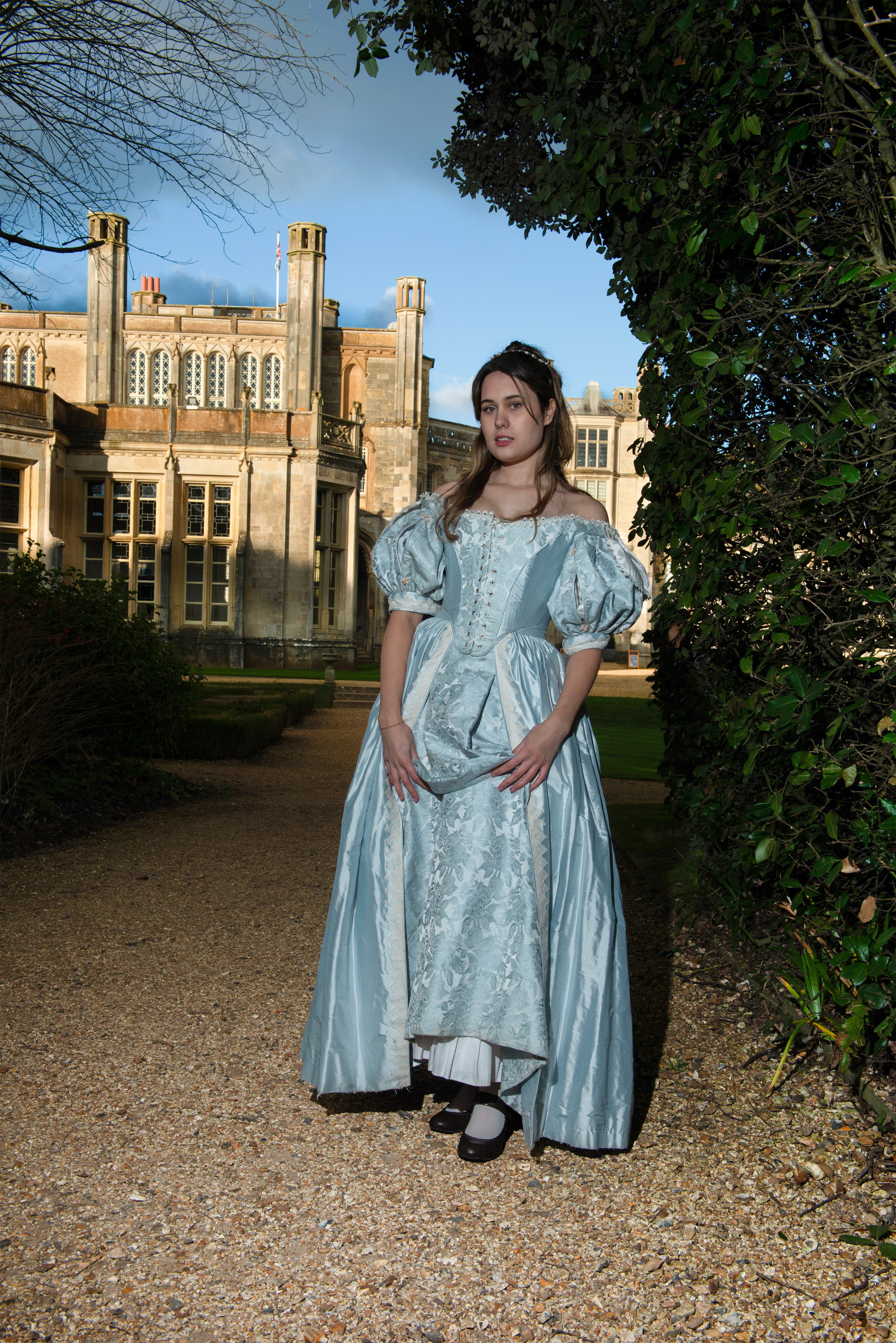 A young woman dressed in a pastel blue historically styled gown standing outdoors on a gravel path with a large historic building in the background, trees at the edges, and clear blue sky.