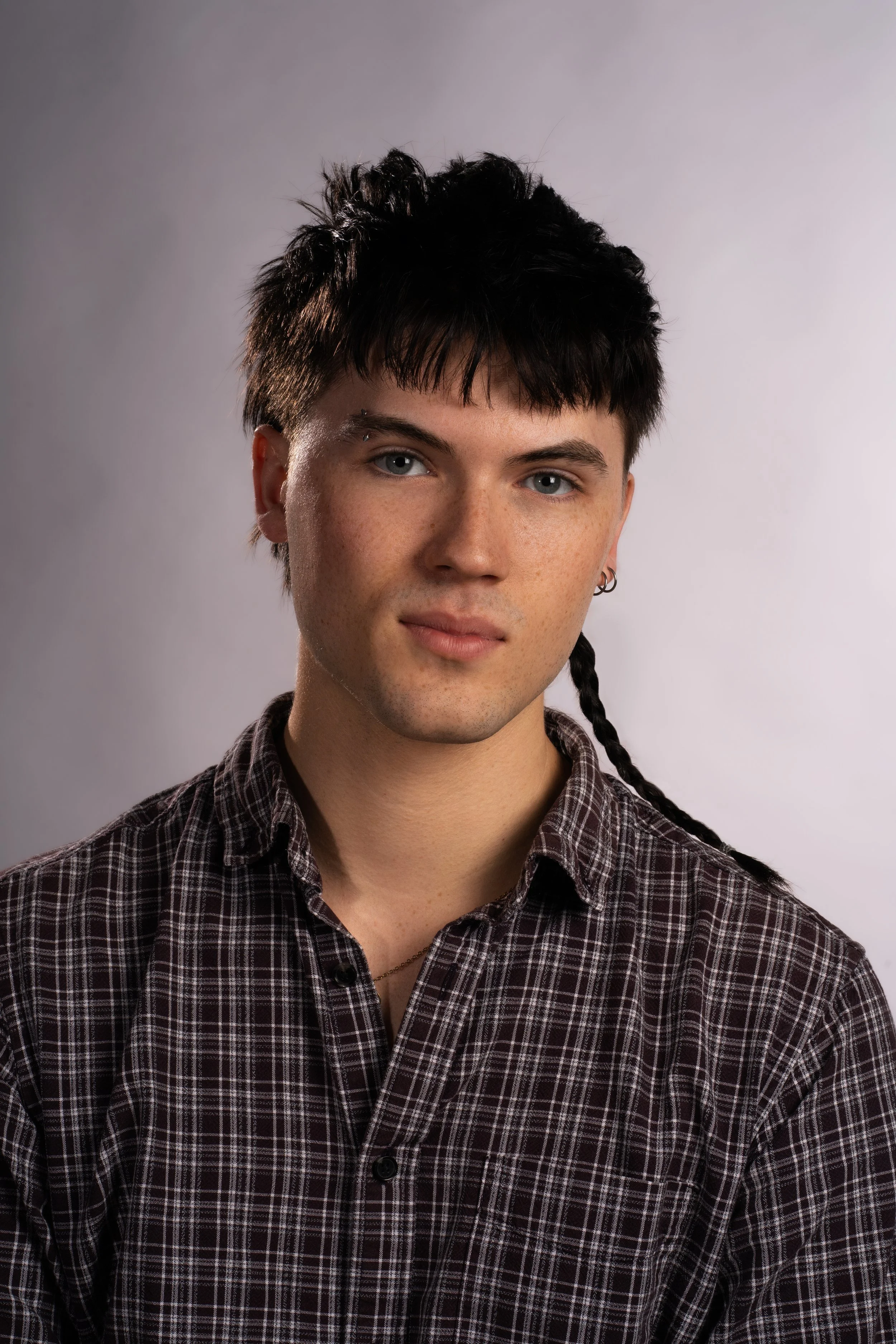 A young person with short, dark, messy hair and blue eyes, wearing a plaid shirt with a collar and a small earring, against a plain light background.