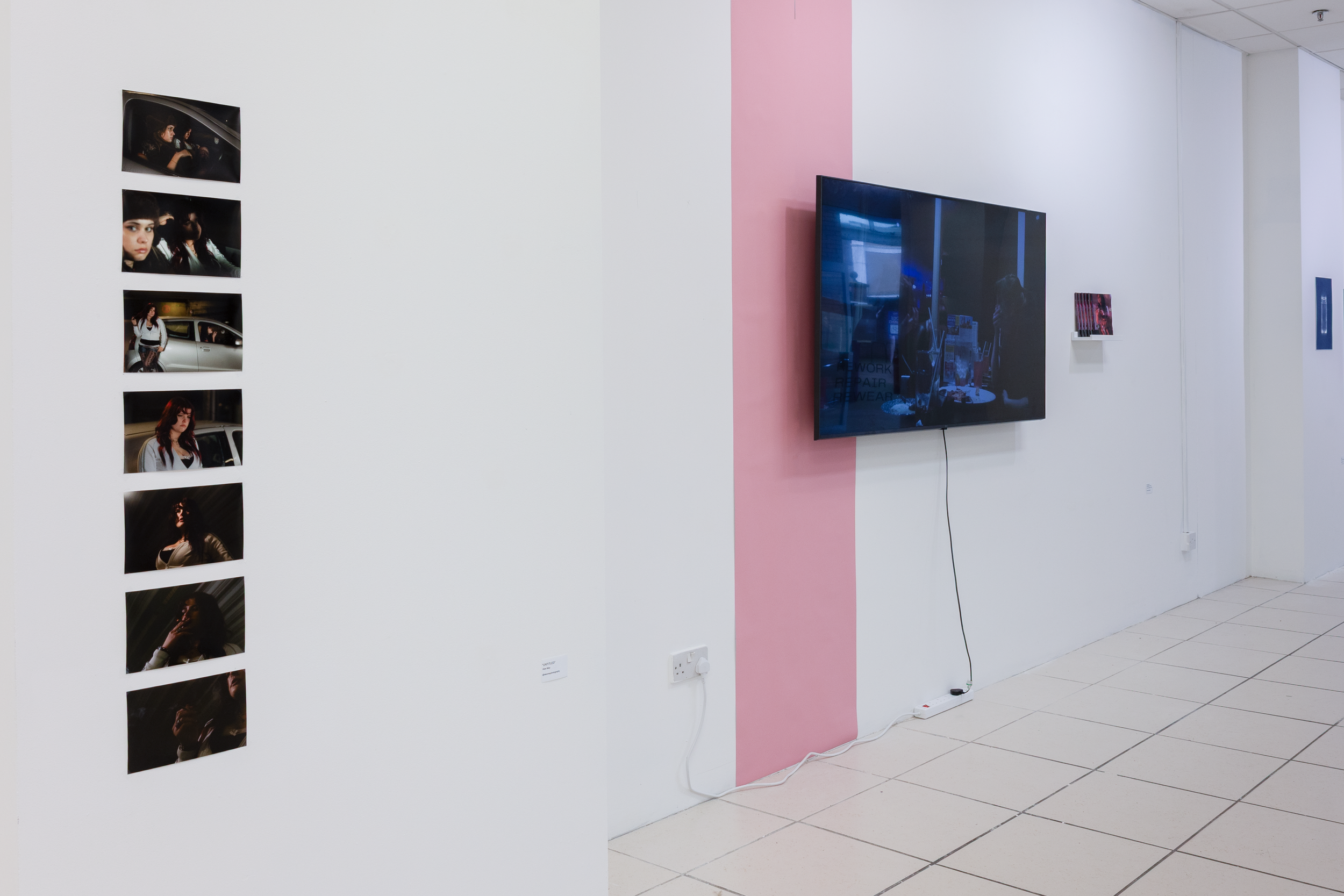 Art gallery with photo series of a woman in different poses and expressions on the left wall, and a large television monitor on the right wall displaying artwork or images, with minimalistic decor and plain white walls.