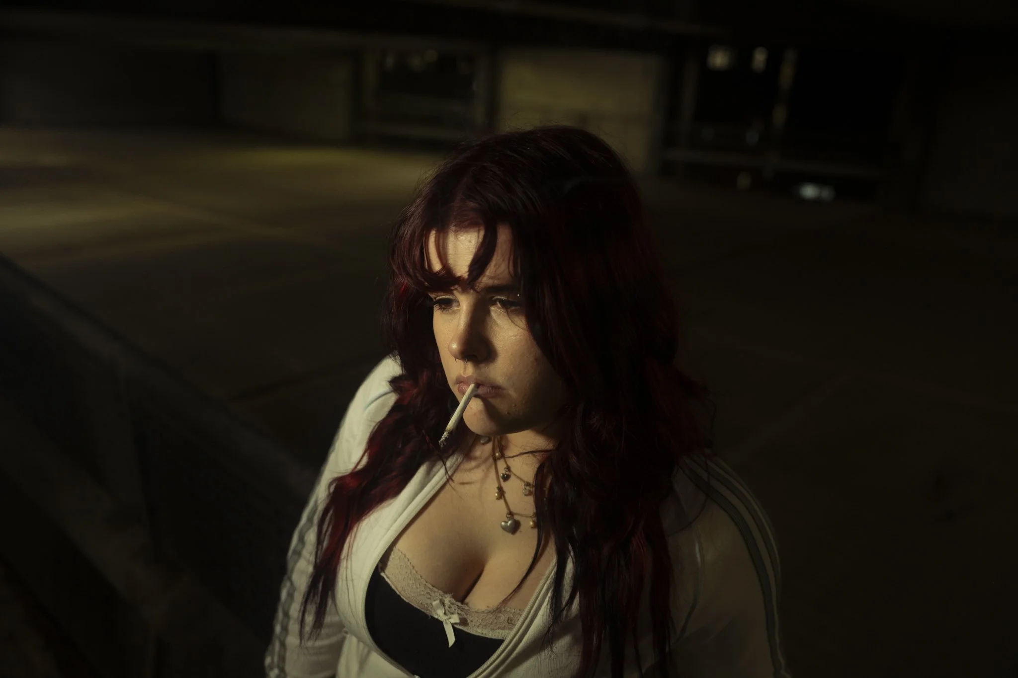 A woman with long dark red hair, holding a cigarette in her mouth, wearing a white jacket over a black top, sitting in a dark, dimly lit room.