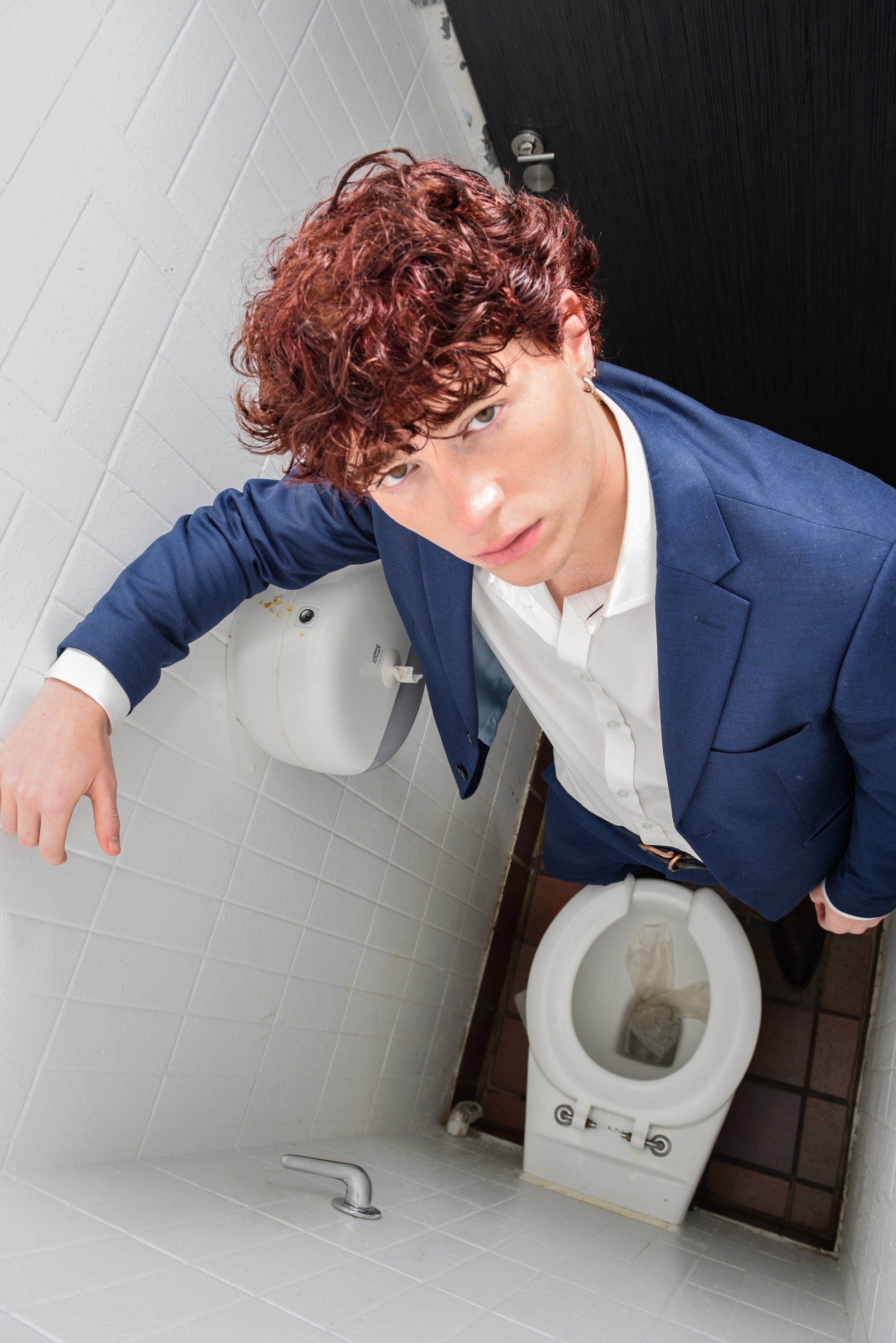 A young man with curly red hair in a blue suit and white shirt appears to be sitting on a closed toilet in a bathroom, looking up at the camera.